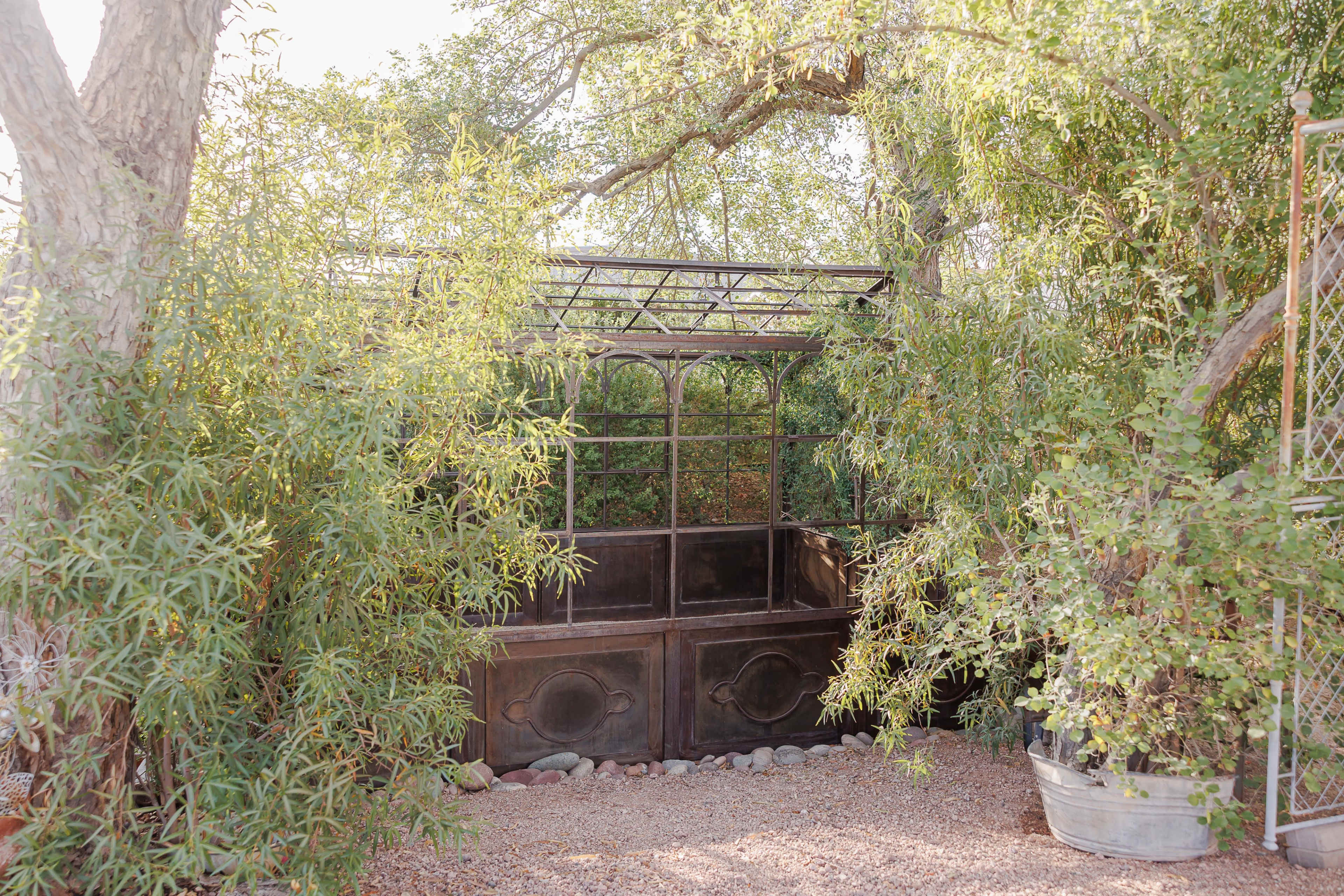 The image shows a greenhouse made of metal and glass, surrounded by lush greenery and rocks on the ground.
