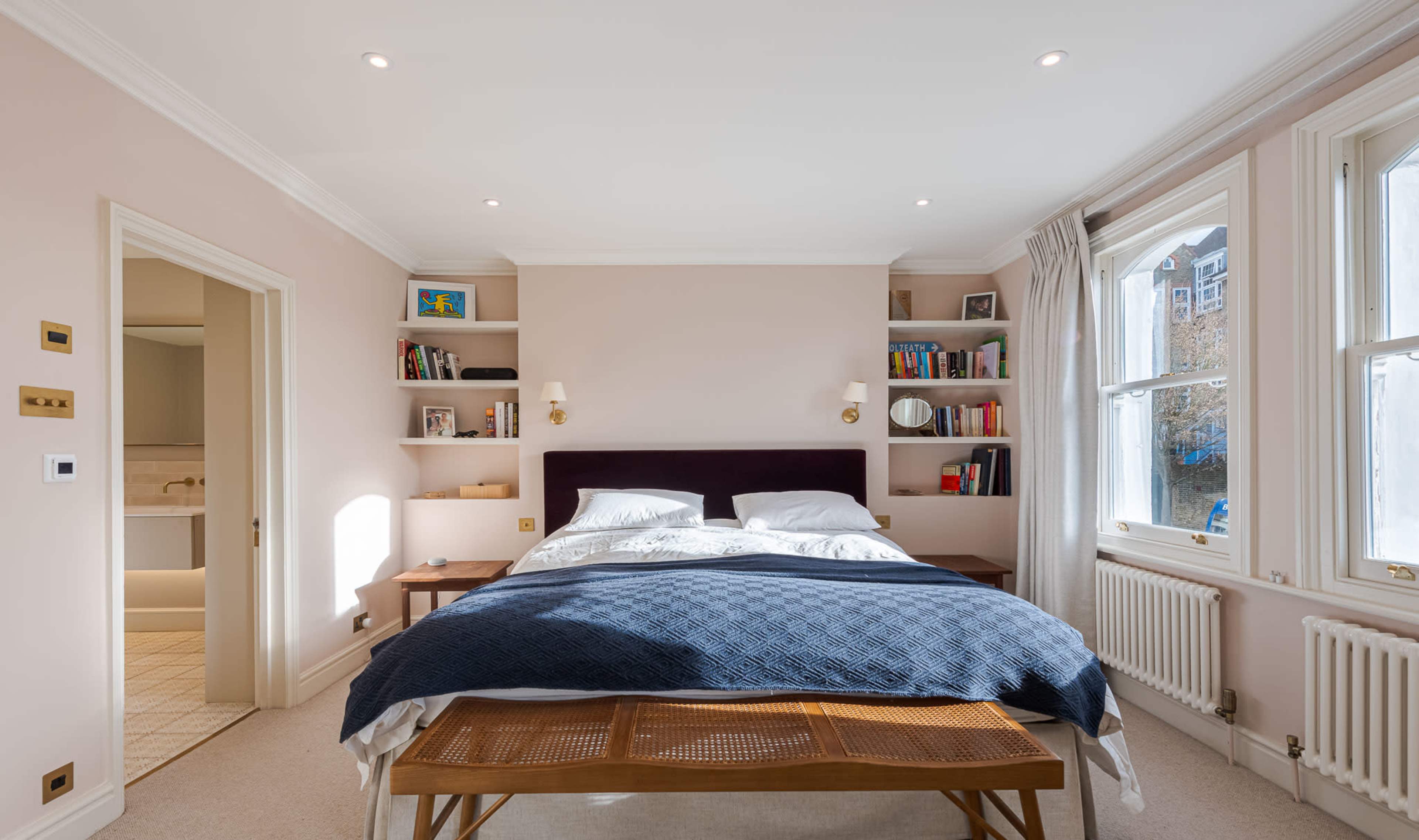 The image shows a well-lit bedroom featuring a double bed with dark bedding, flanked by shelves with books and a small side table.