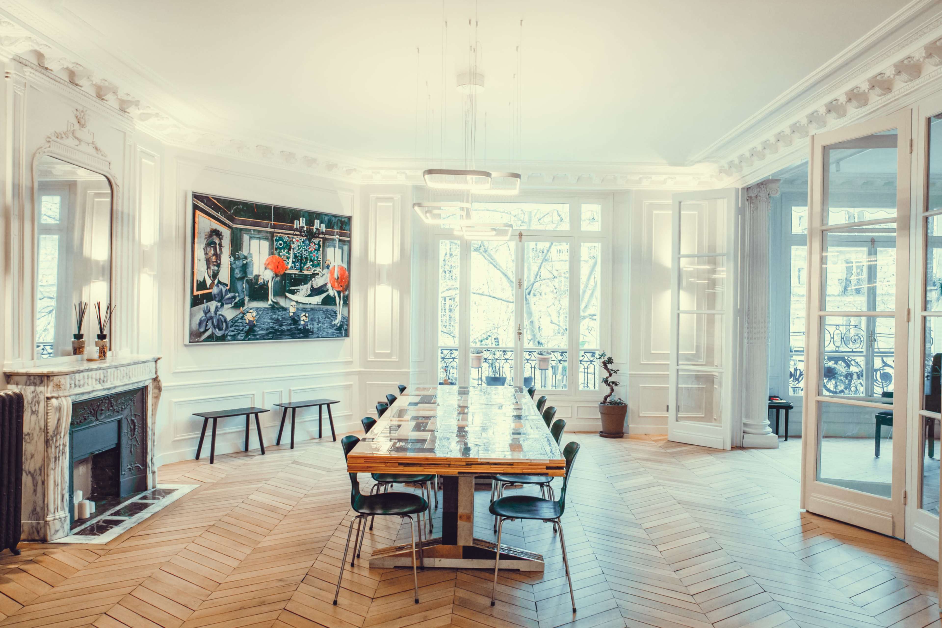 The image shows a well-lit dining room with a large wooden table, black chairs, and a decorative fireplace, featuring an artwork on the wall and large windows overlooking a balcony.