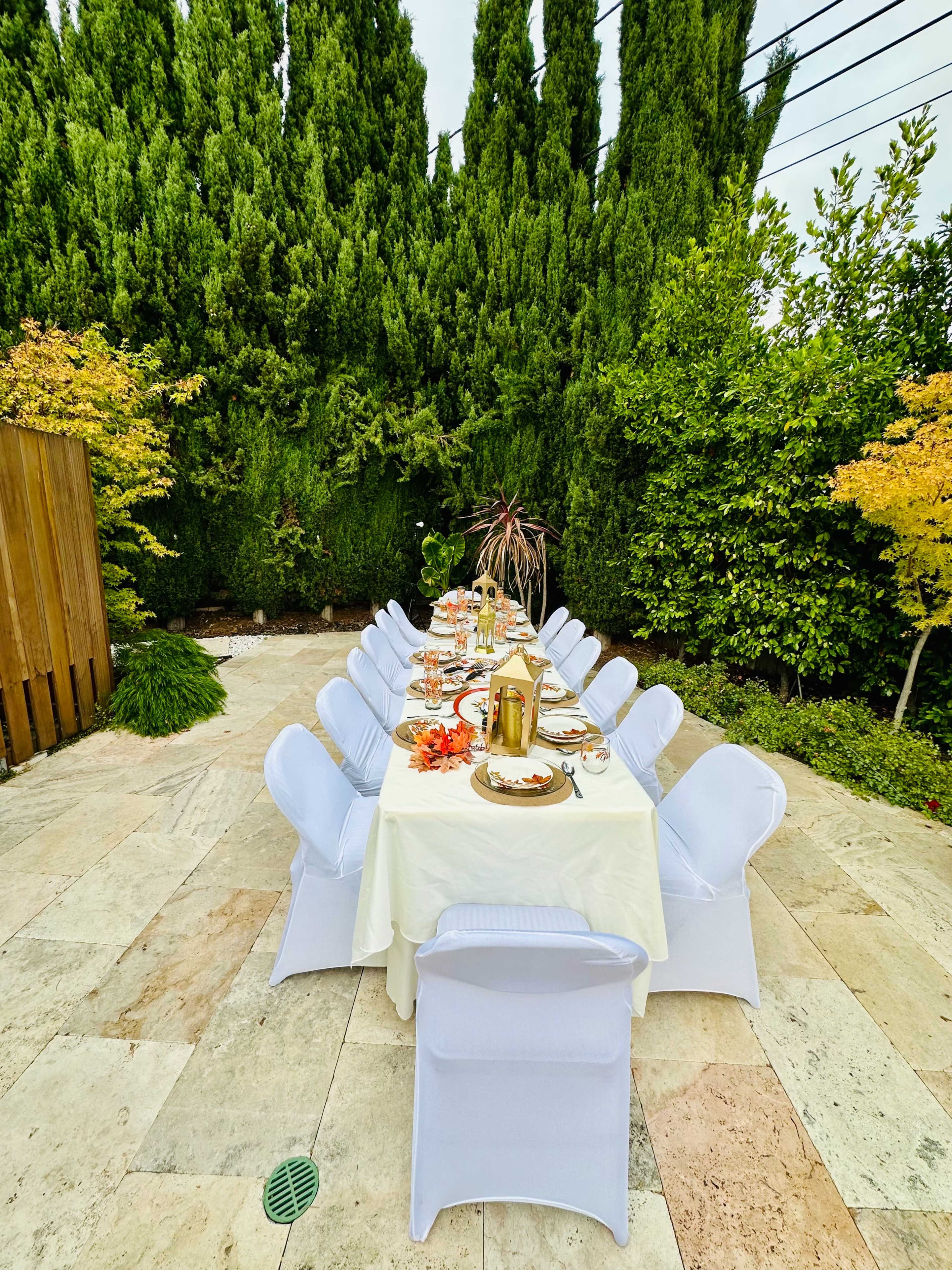 A long dining table is set outdoors with white chairs and colorful autumn decorations, surrounded by tall greenery.