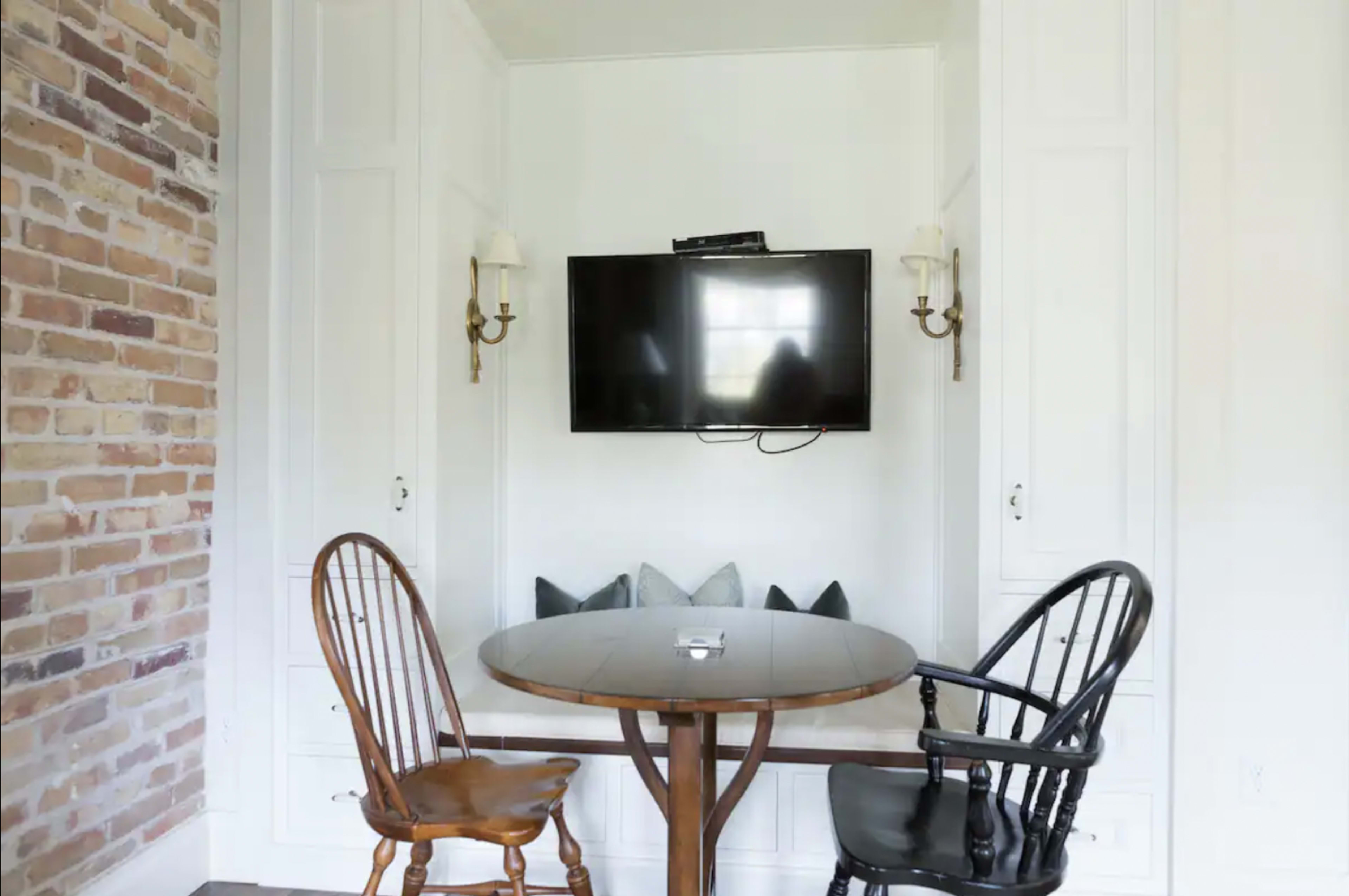 A small dining area with a round table and two chairs, one wooden and one black, positioned against a wall with a television mounted above and a cozy nook behind the table.