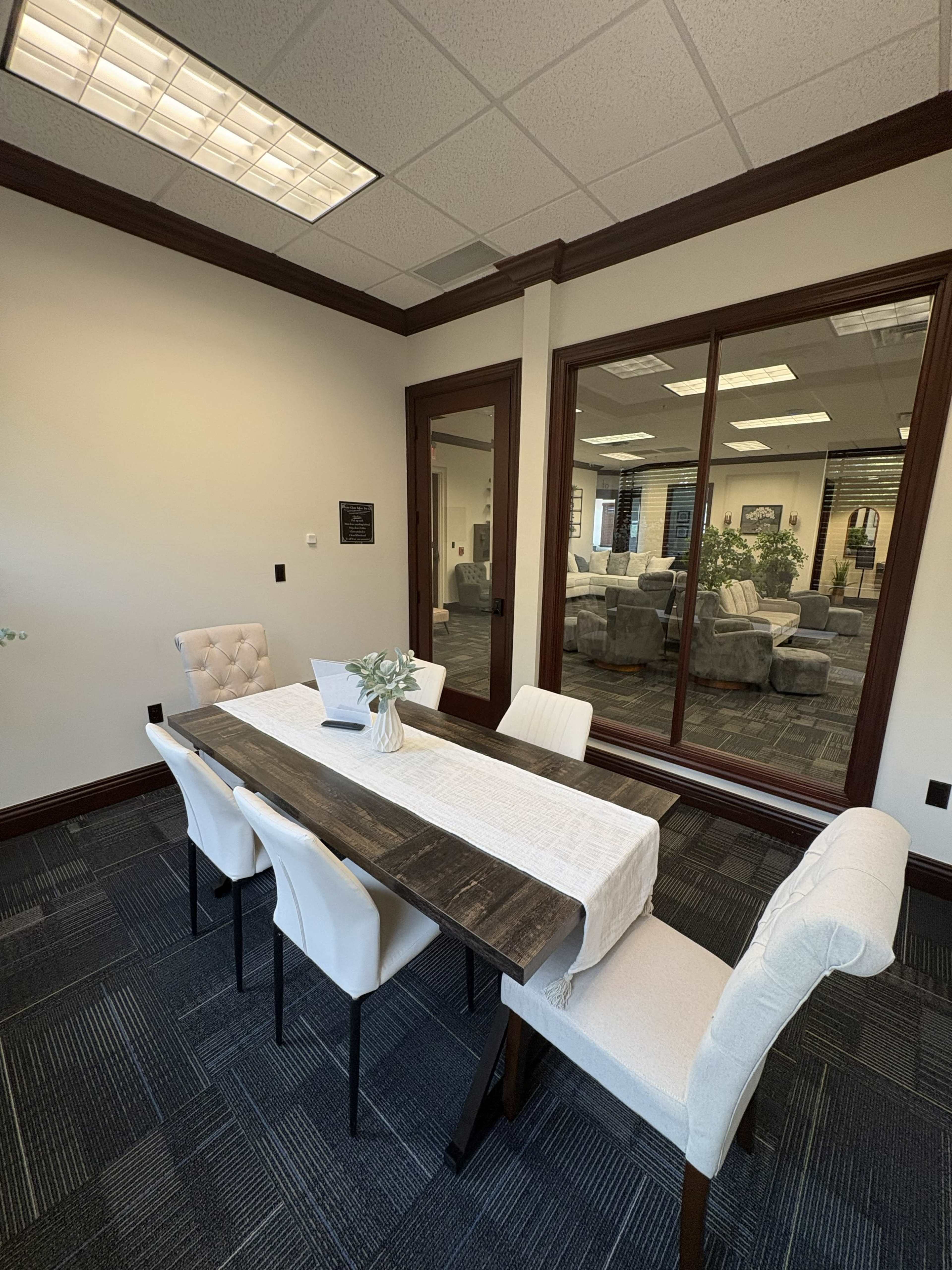 A conference room features a wooden table with a white table runner and four upholstered chairs, with large windows reflecting a spacious office area.