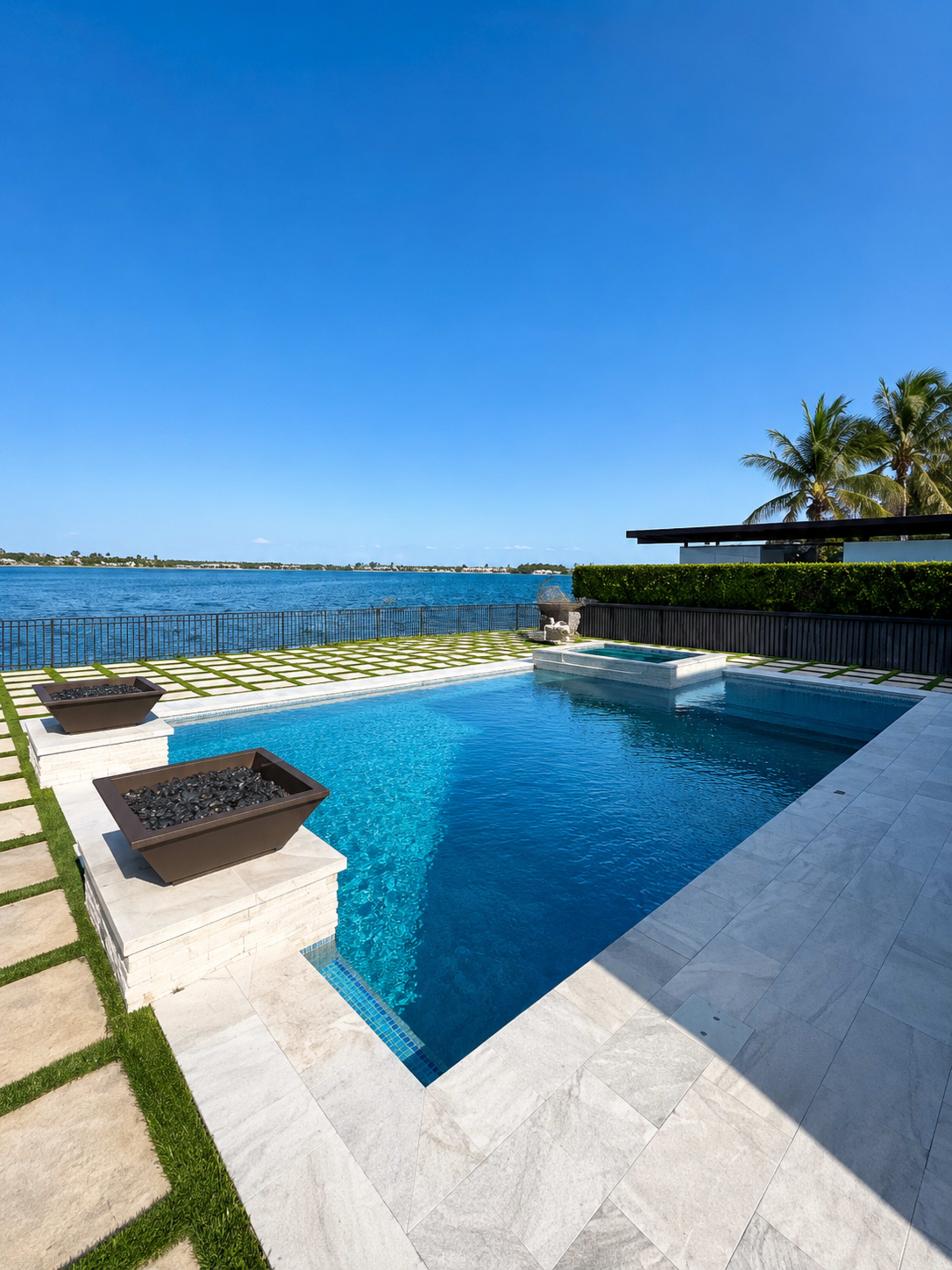 The image shows a modern swimming pool with a stone patio, surrounded by manicured grass and overlooking a body of water.