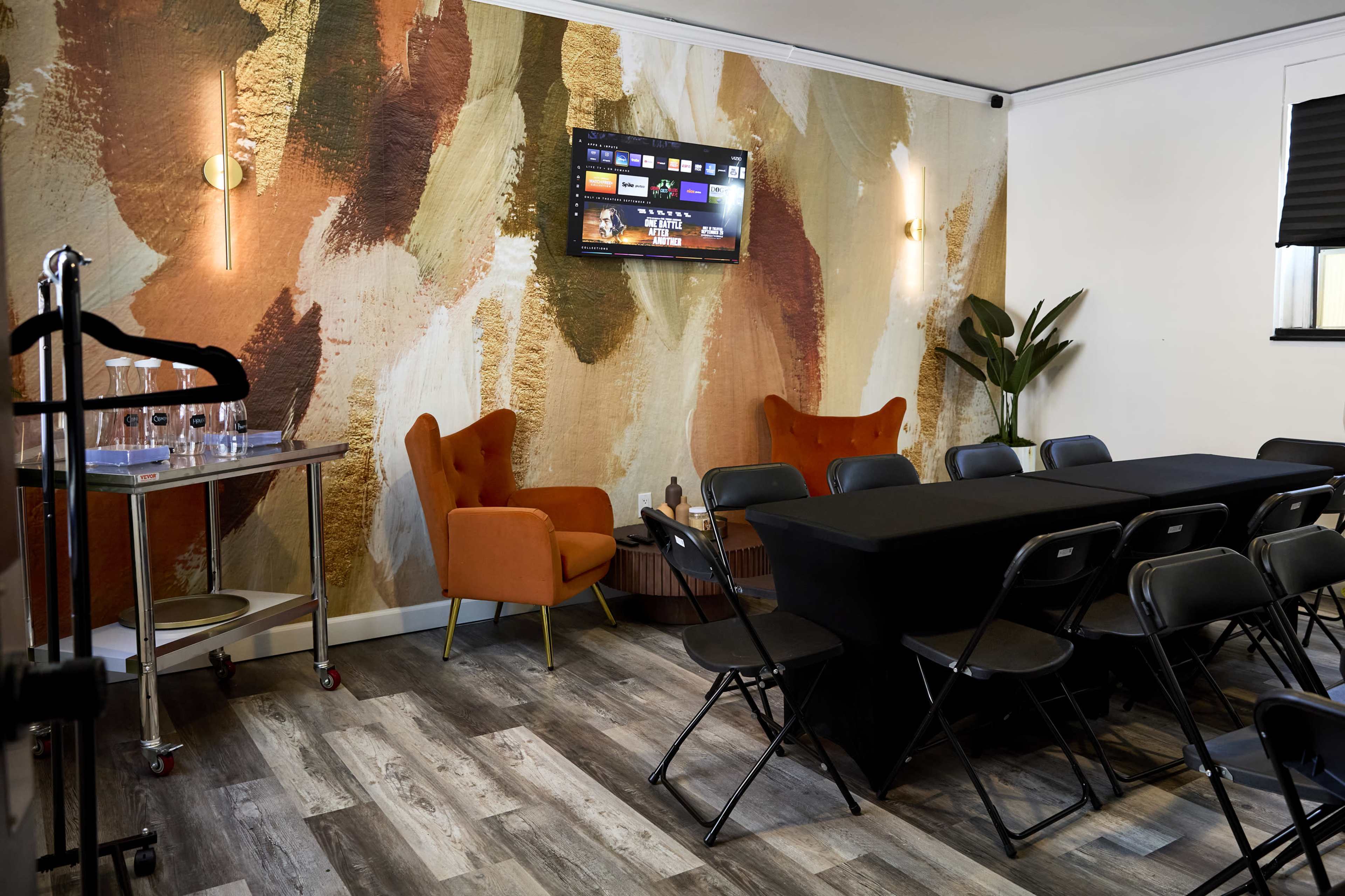 A meeting room features a screen mounted on a textured wall, with orange armchairs, a black conference table, and rows of black folding chairs.