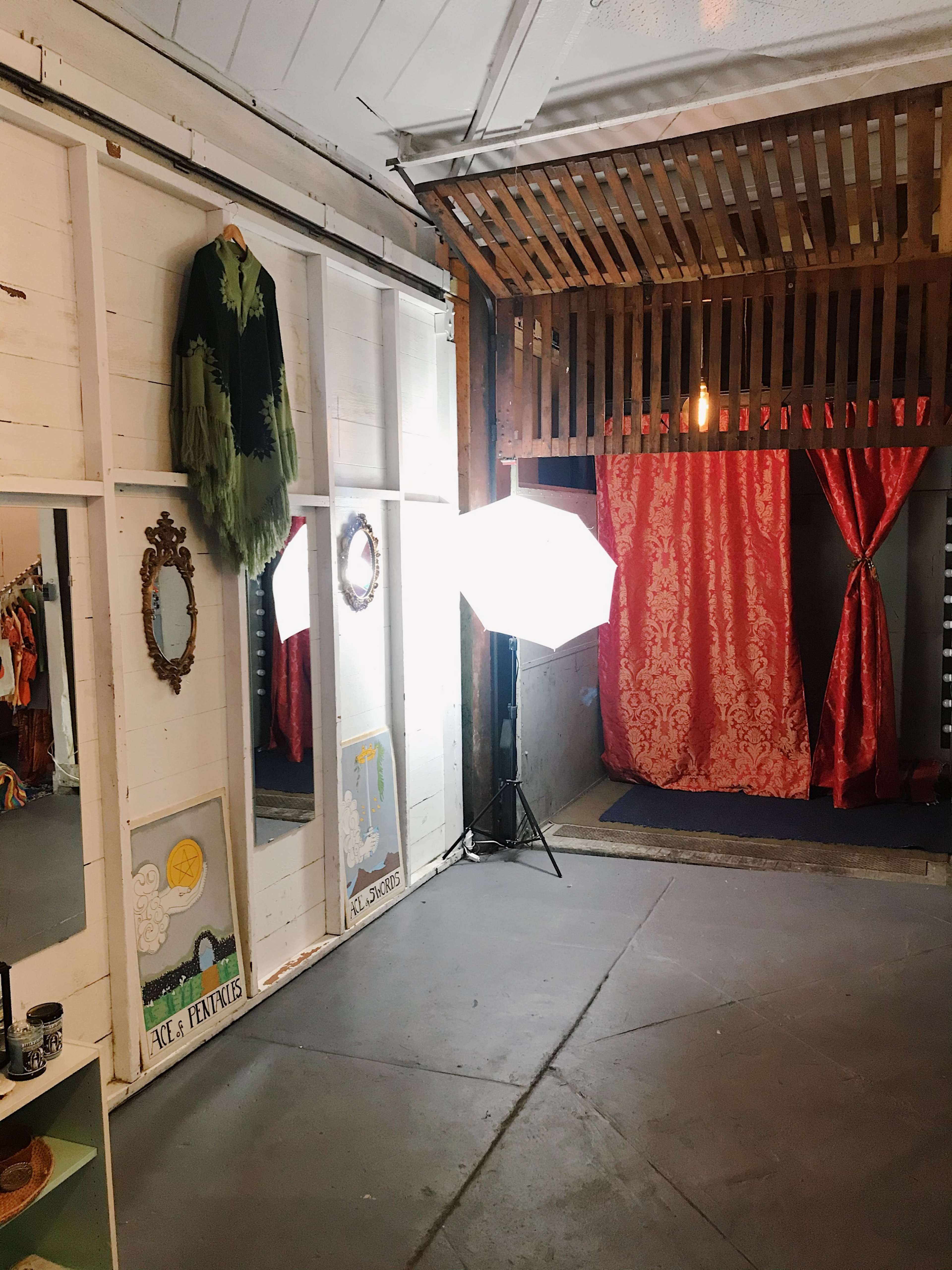 A room with mirrors, a garment hanging on the wall, and a backdrop of red curtains, illuminated by a softbox light.