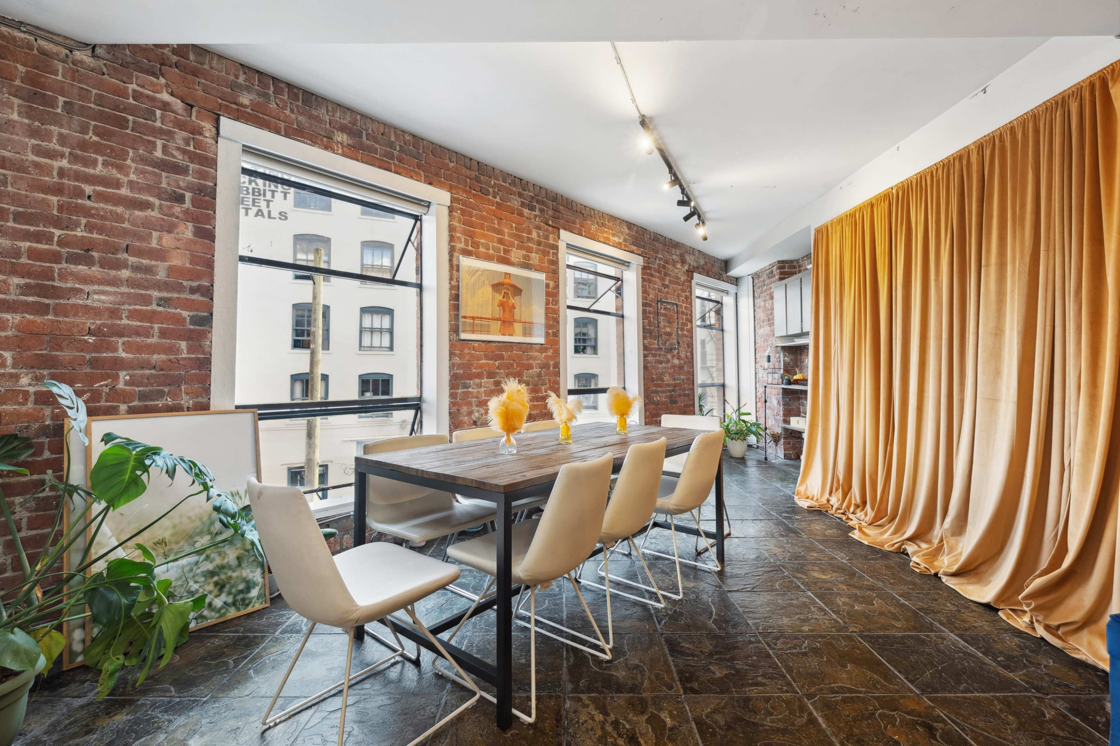 A dining area with a wooden table and six white chairs, surrounded by exposed brick walls and large windows, with a yellow curtain draped in the corner.
