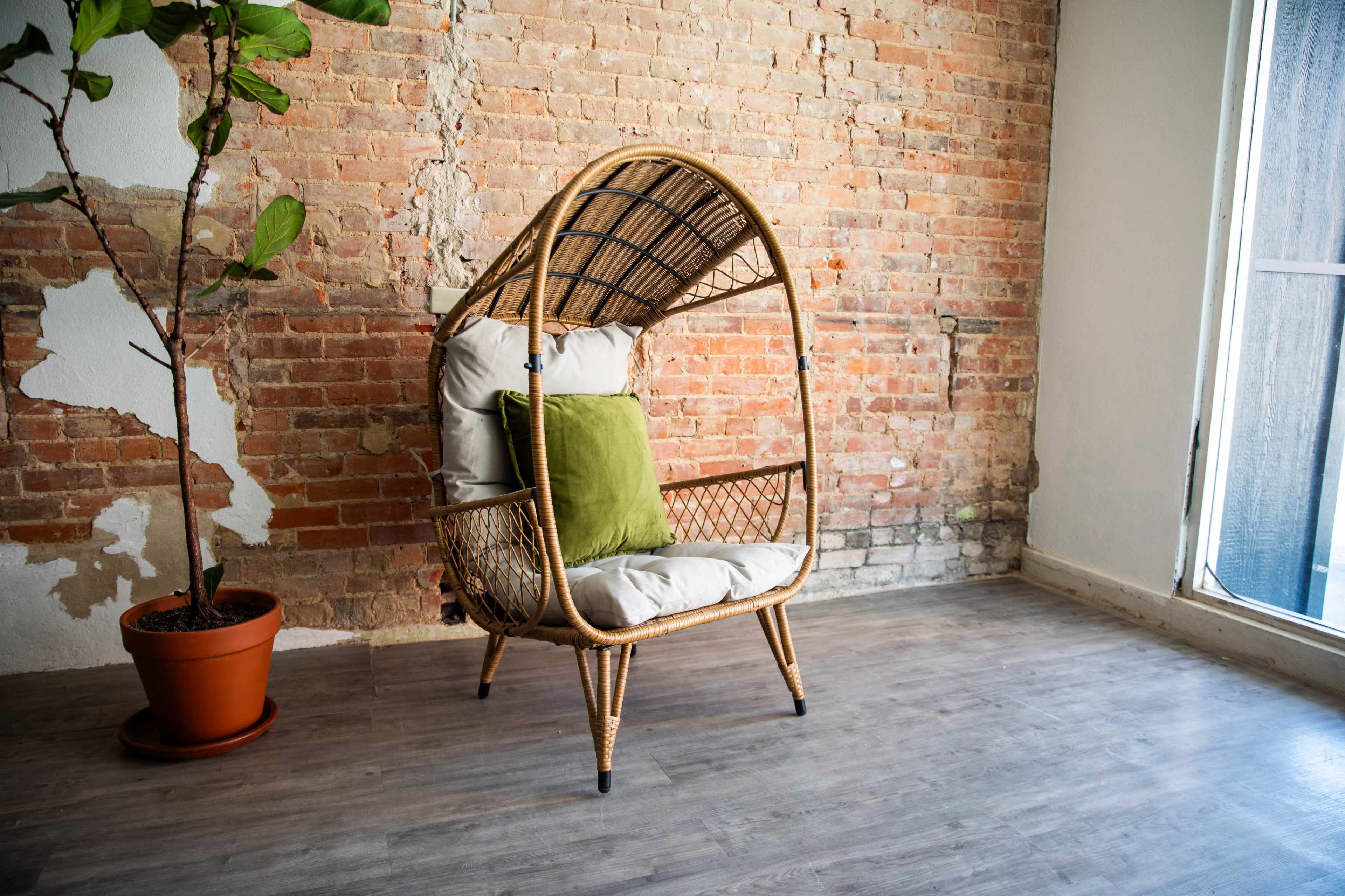 A rattan chair with a curved canopy is positioned against a brick wall, accompanied by a potted plant.