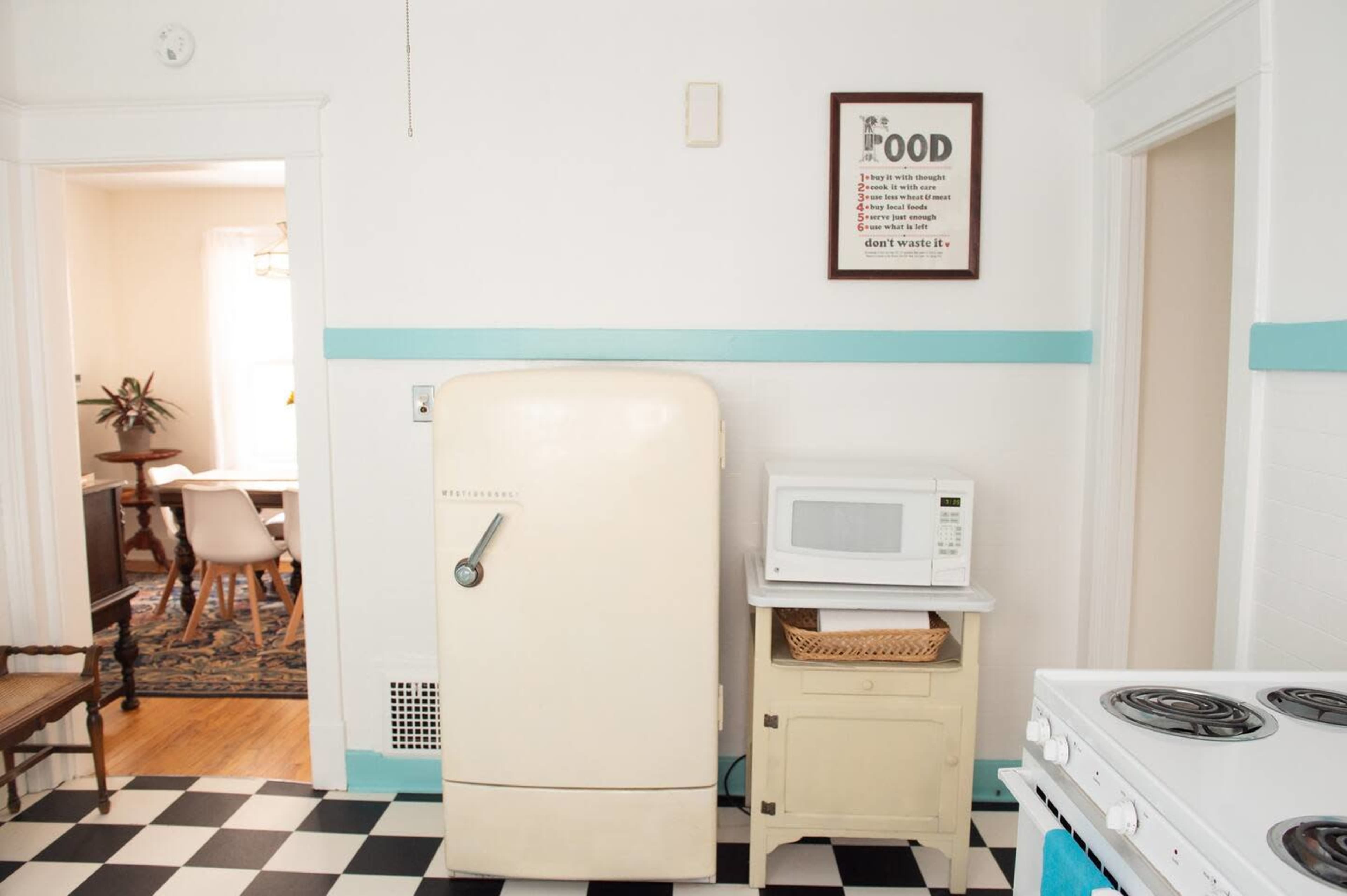 A vintage refrigerator stands next to a microwave in a kitchen with black and white checkered flooring and a doorway leading to a dining area.