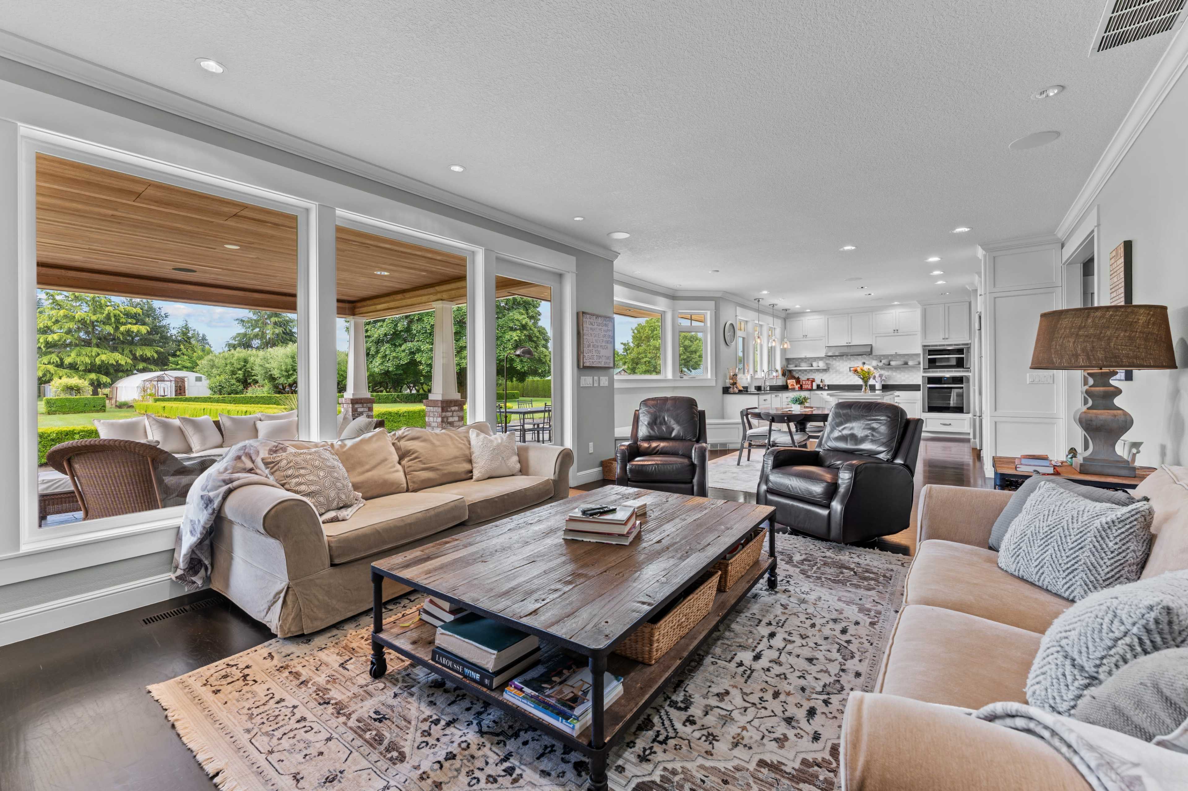 The living room features a spacious layout with large windows, a coffee table at the center, and seating arrangements that include a beige sofa and dark leather chairs, all overlooking a green outdoor space.