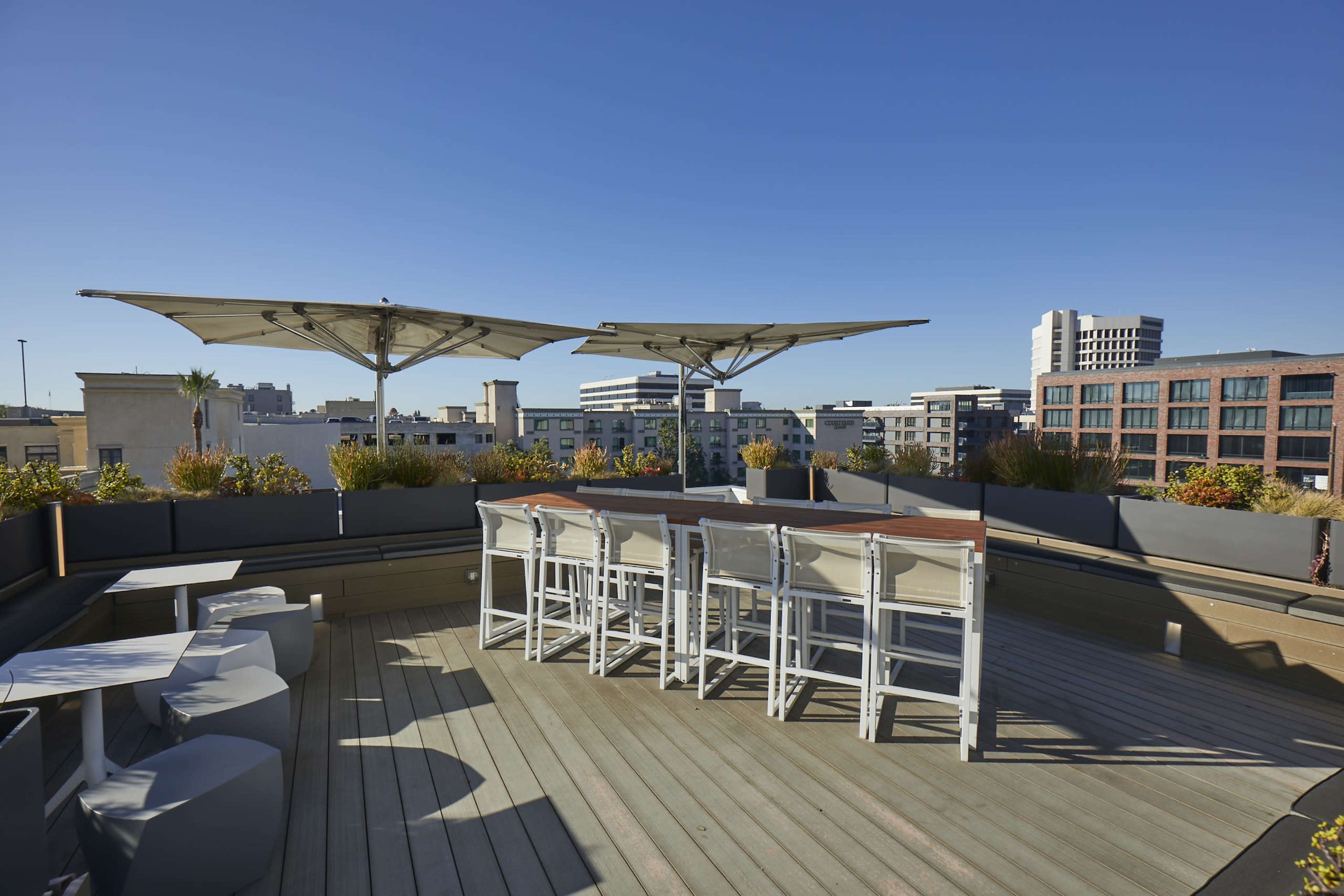 A spacious rooftop area with a wooden deck, a bar table and chairs, and large shading canopies overhead, surrounded by potted plants.
