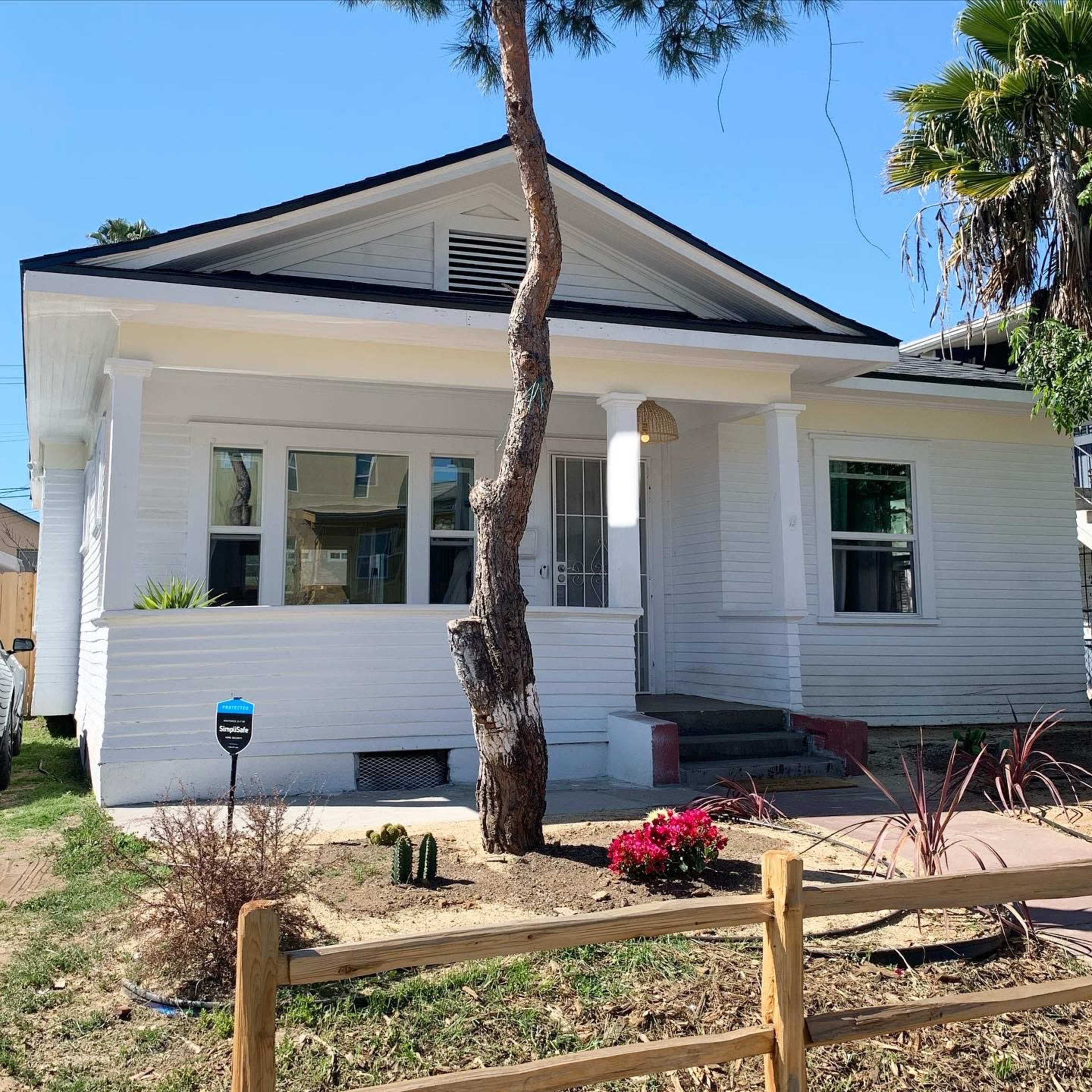 A white, single-story house with a front porch, a slanted roof, and various plants in the yard.