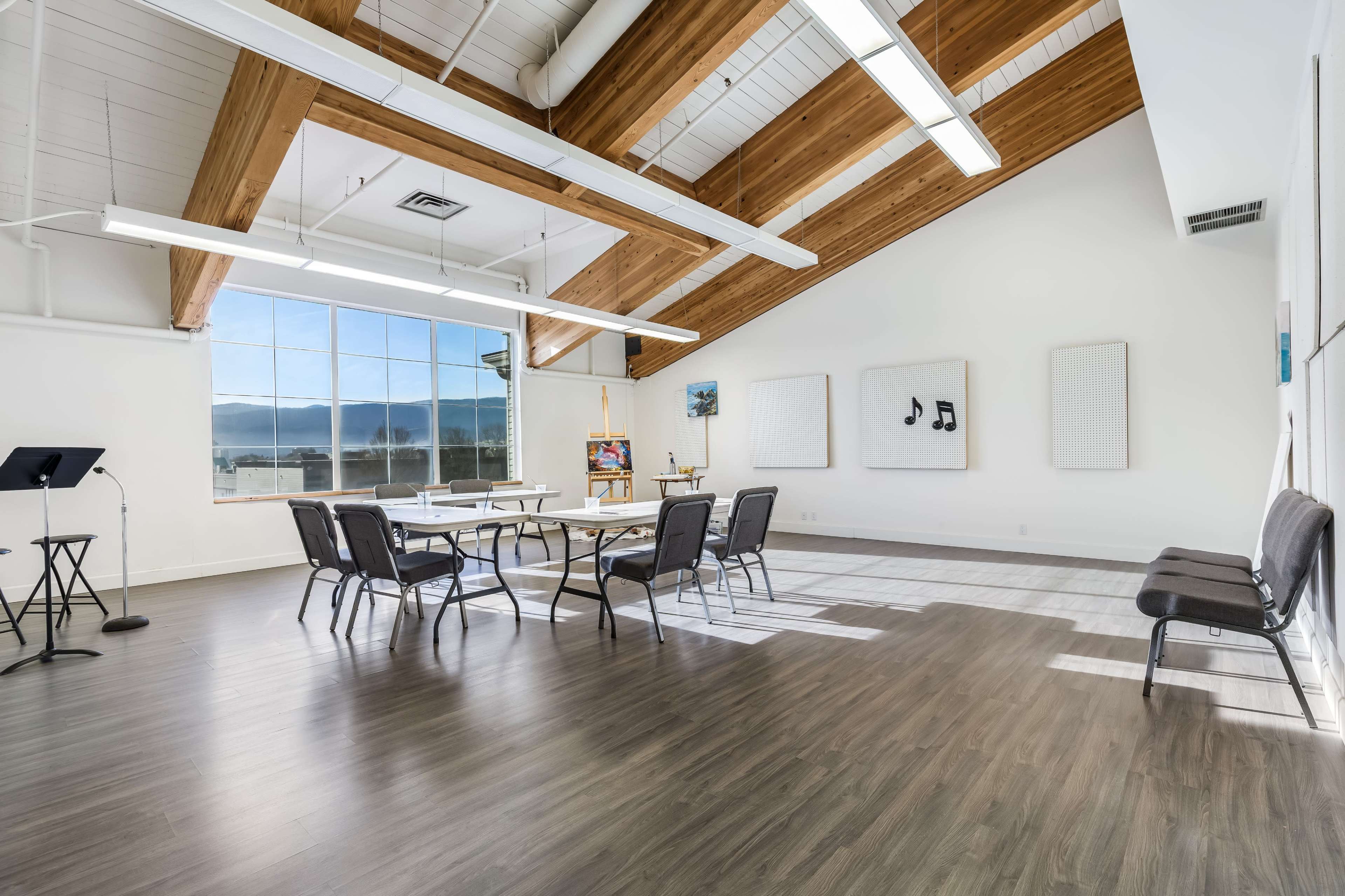 The image shows a spacious, well-lit room with high ceilings, wooden beams, several tables and chairs arranged, and large windows with a view of mountains in the background.