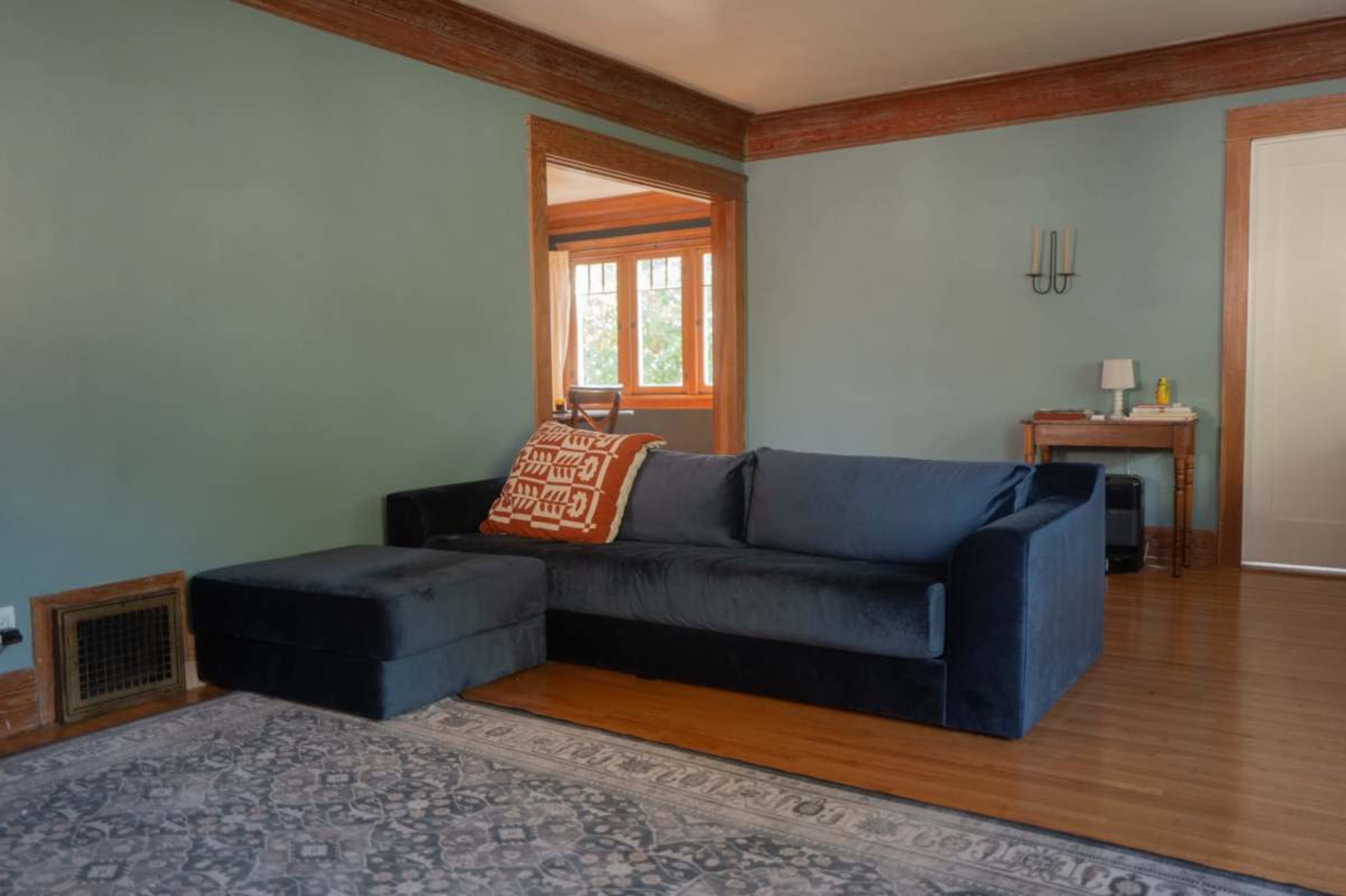 A dark blue sectional sofa with an orange patterned throw pillow is positioned against a wall in a room with light green paint and wooden trim.
