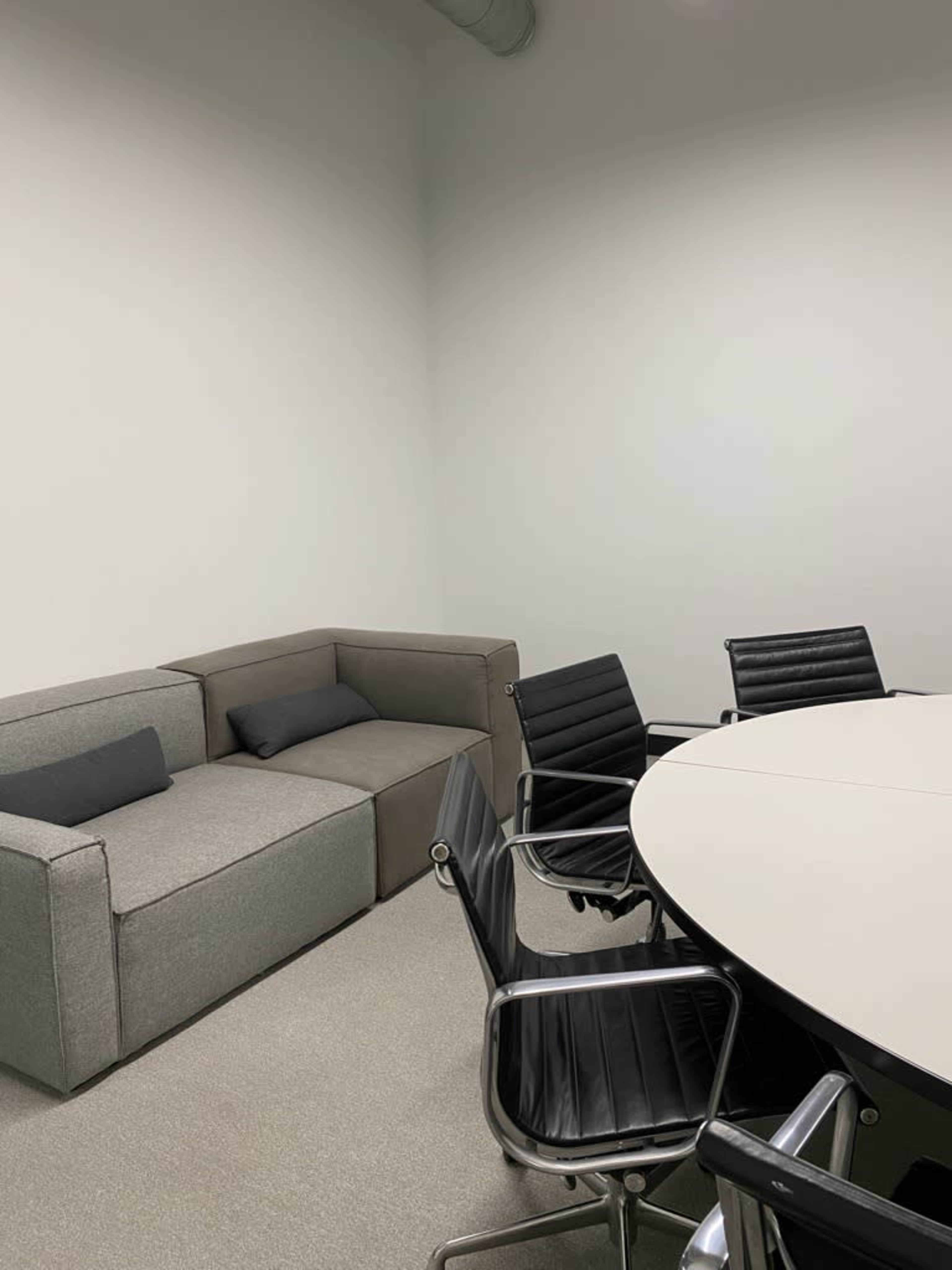 The image shows a small meeting room with a round table surrounded by black chairs and a gray sectional couch in one corner.