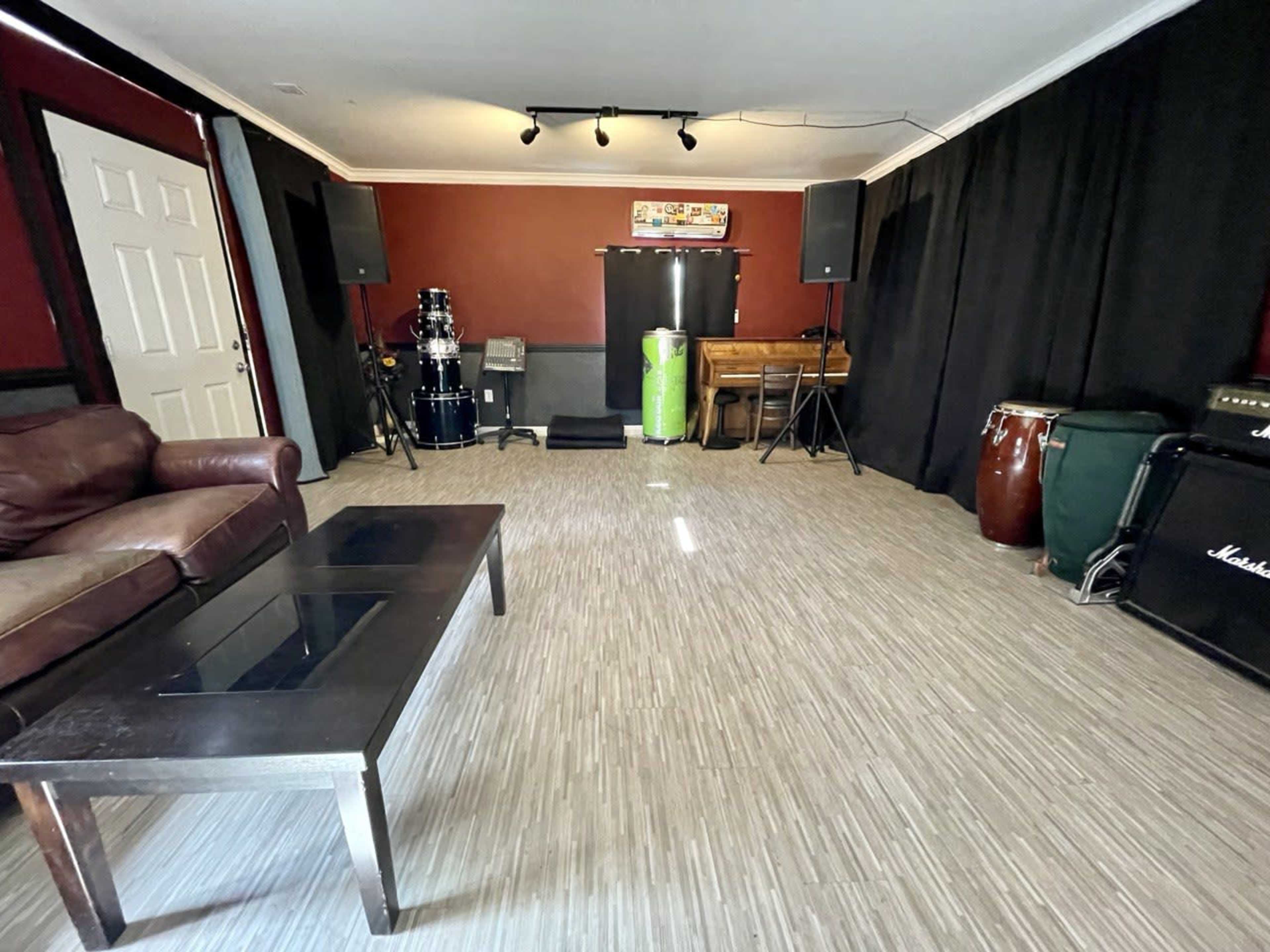 A music rehearsal space featuring a brown leather couch, a coffee table, various musical instruments, and sound equipment against a backdrop of black curtains.