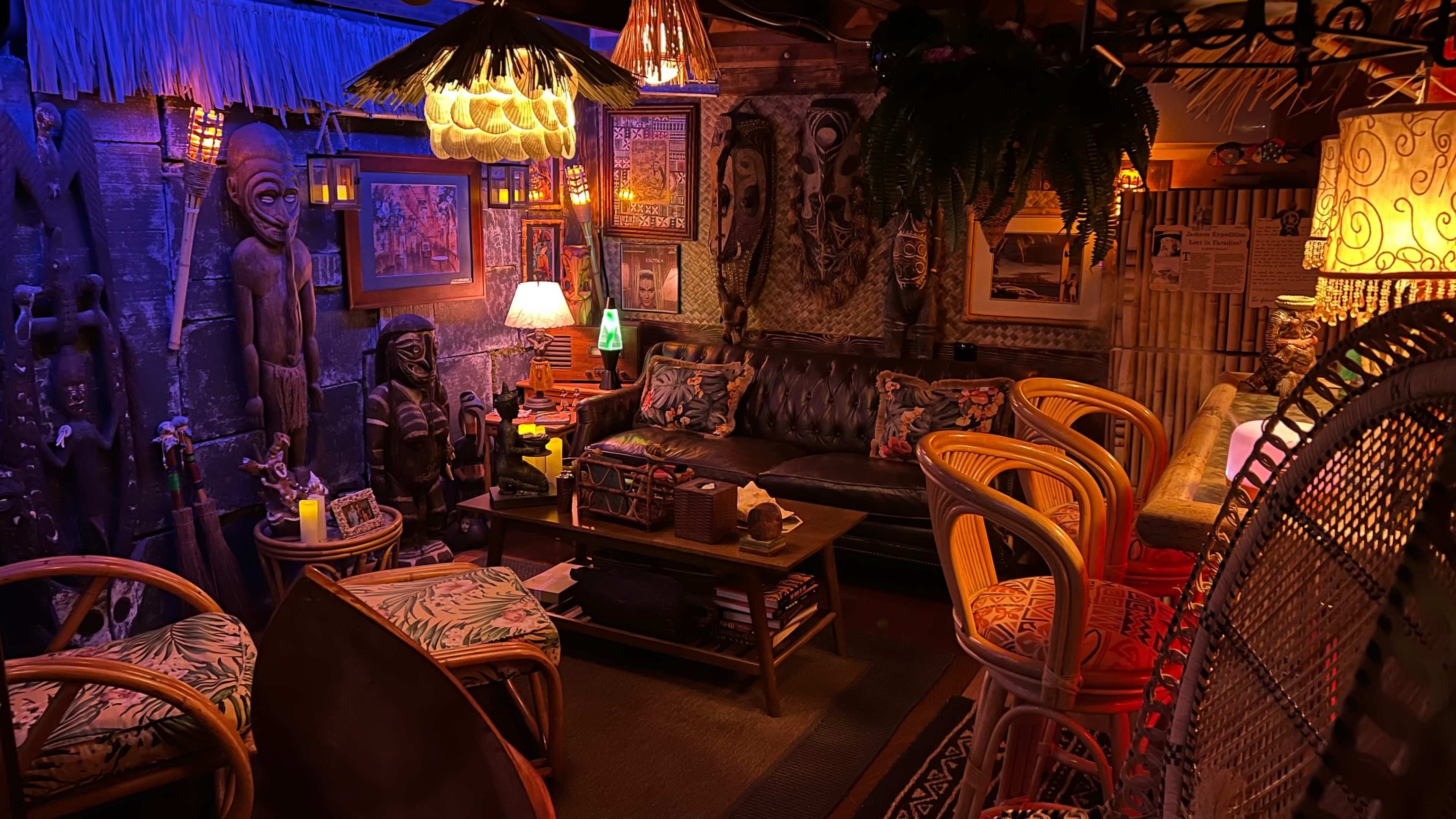 The image shows a dimly lit, decorated lounge area featuring a leather sofa, bamboo chairs, and various artworks and sculptures on the walls.