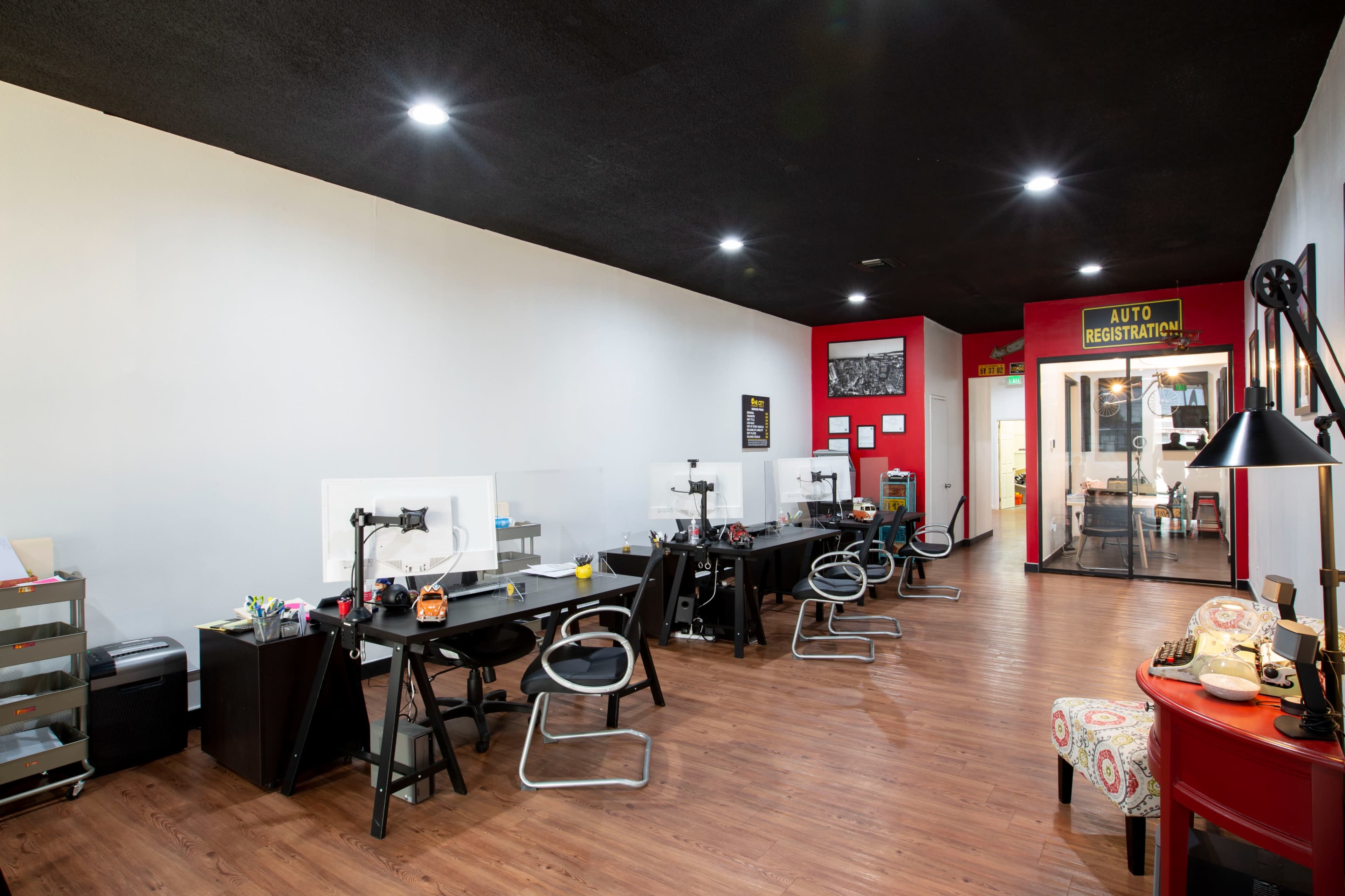 A modern office space with several black desks arranged in rows, equipped with computers and protective partitions, alongside a glass-walled room labeled "AUTO REGISTRATION."