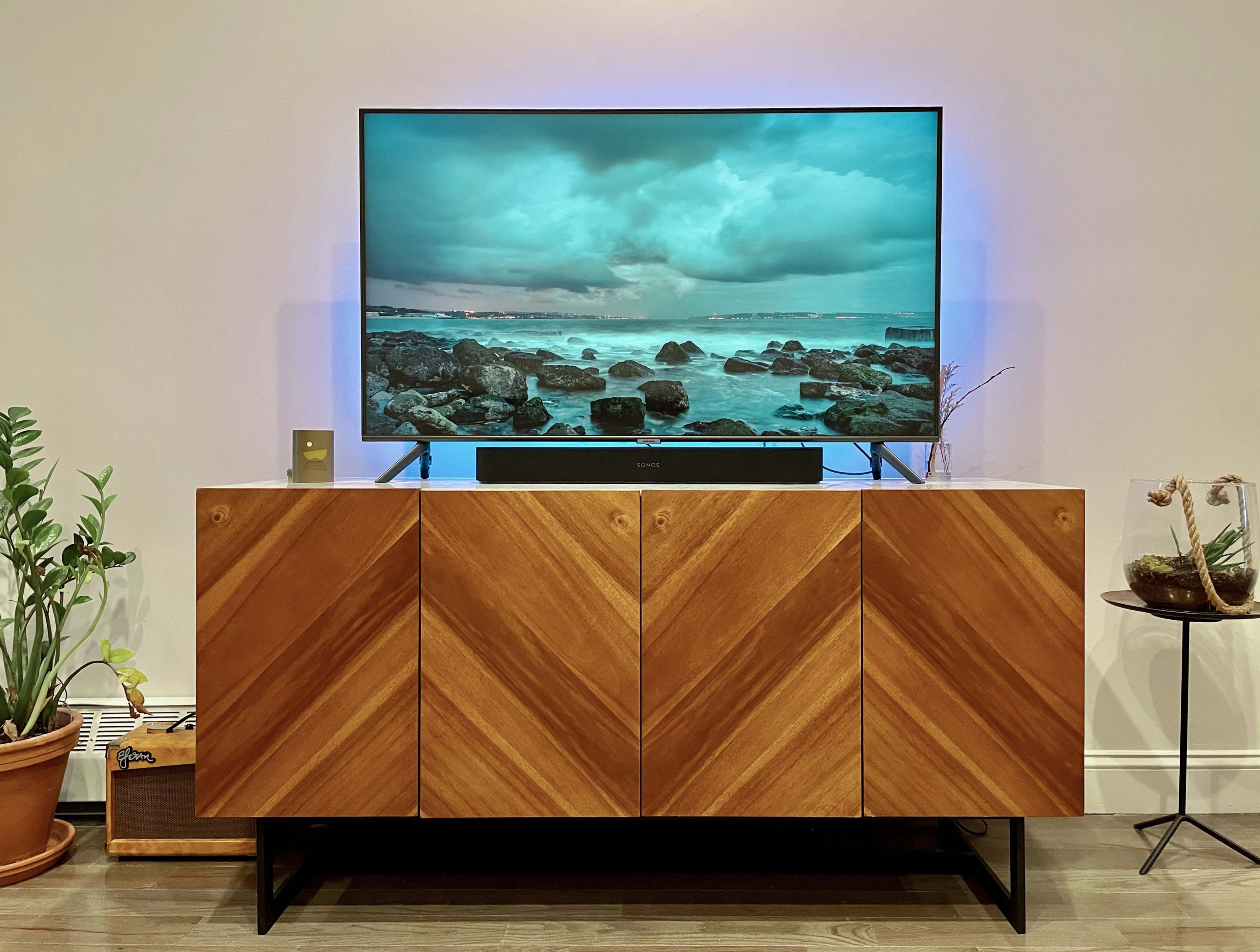 A television displaying a coastal scene is mounted above a wooden cabinet with a chevron pattern, accompanied by potted plants and a small side table.