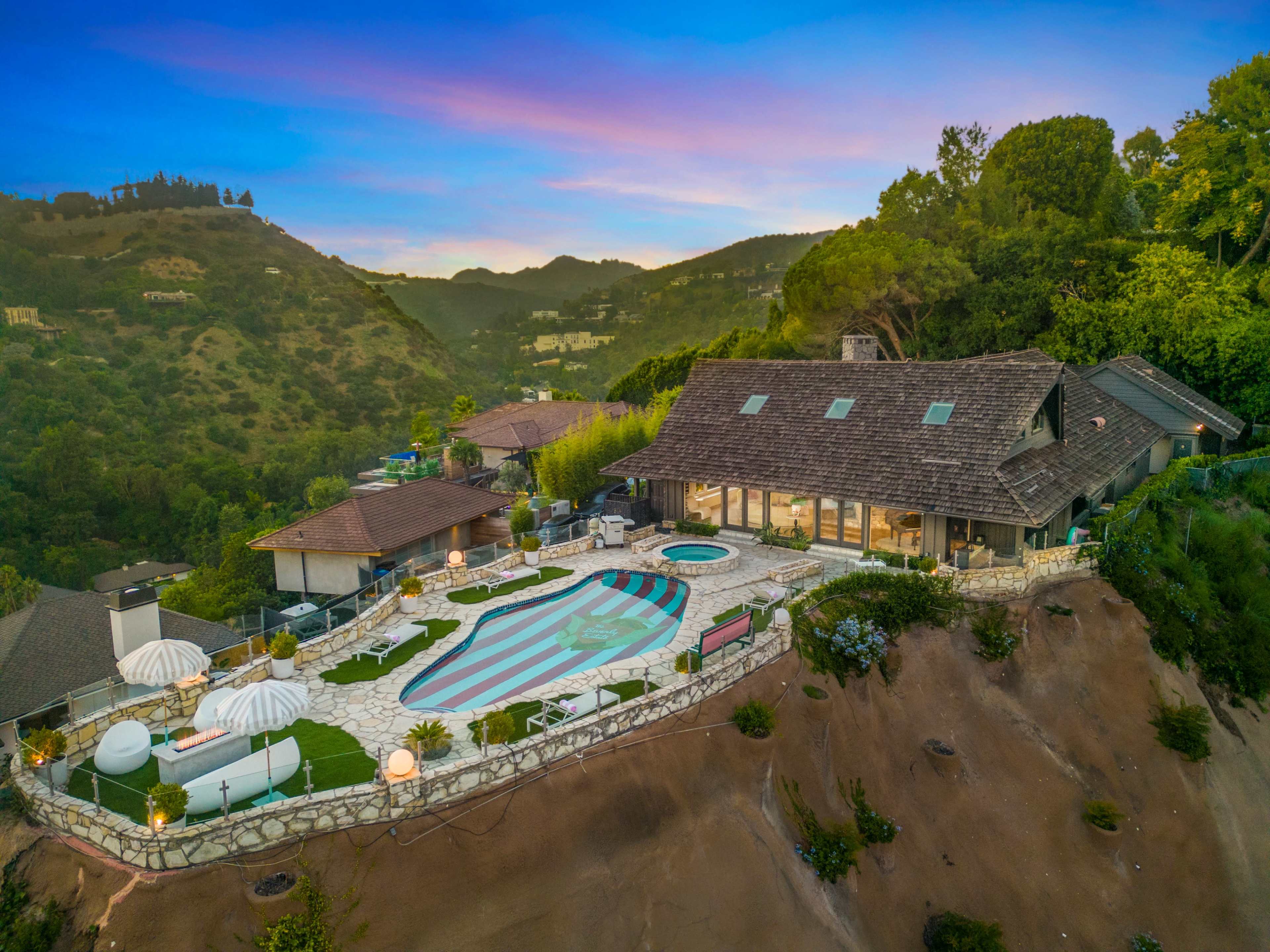 The image depicts a large hillside home with an expansive pool area, surrounded by greenery and offering panoramic views of the surrounding hills at sunset.