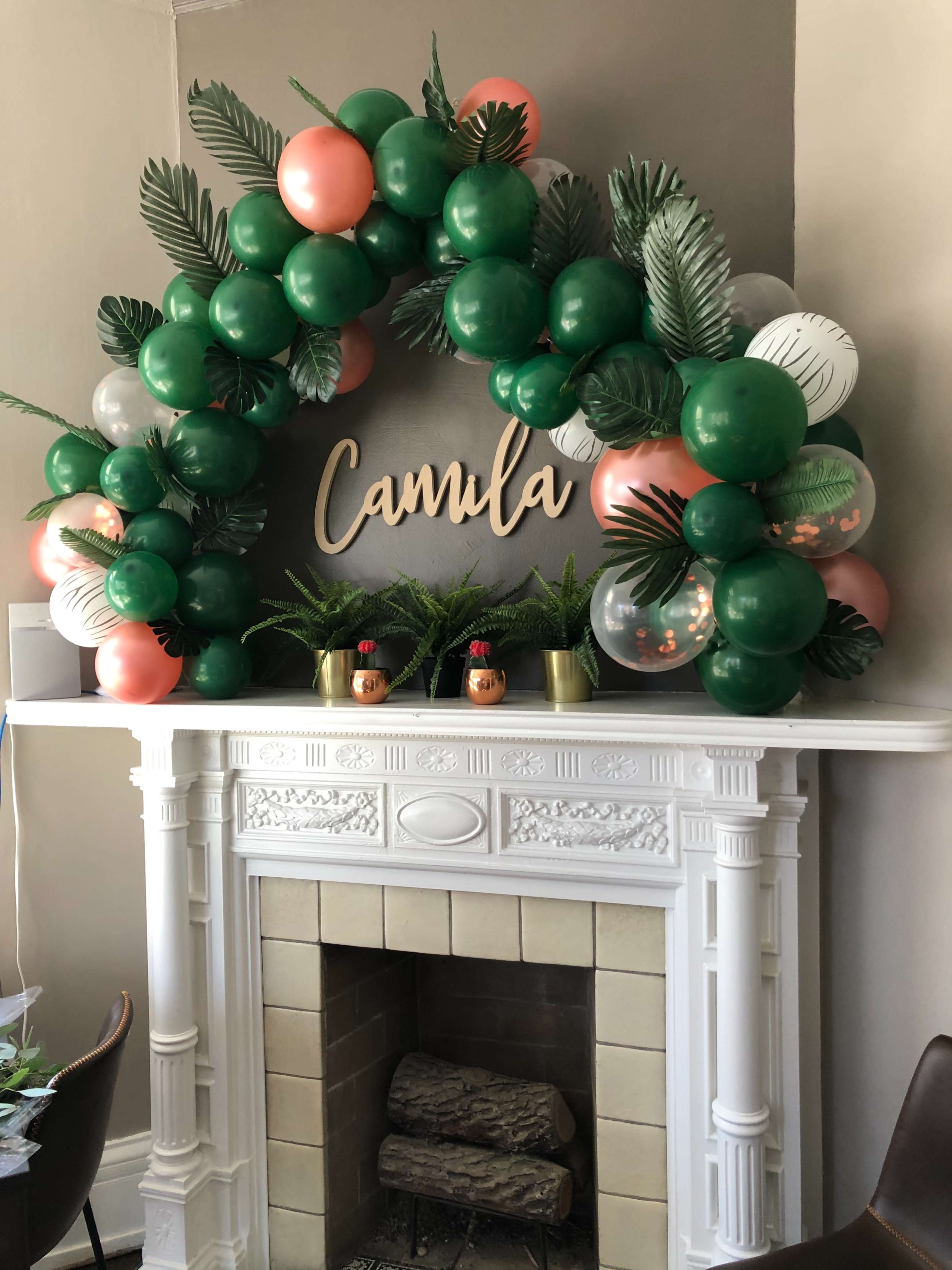 A decorative balloon arch featuring green, pink, and clear balloons is displayed above a fireplace with the name "Camila" and potted ferns.