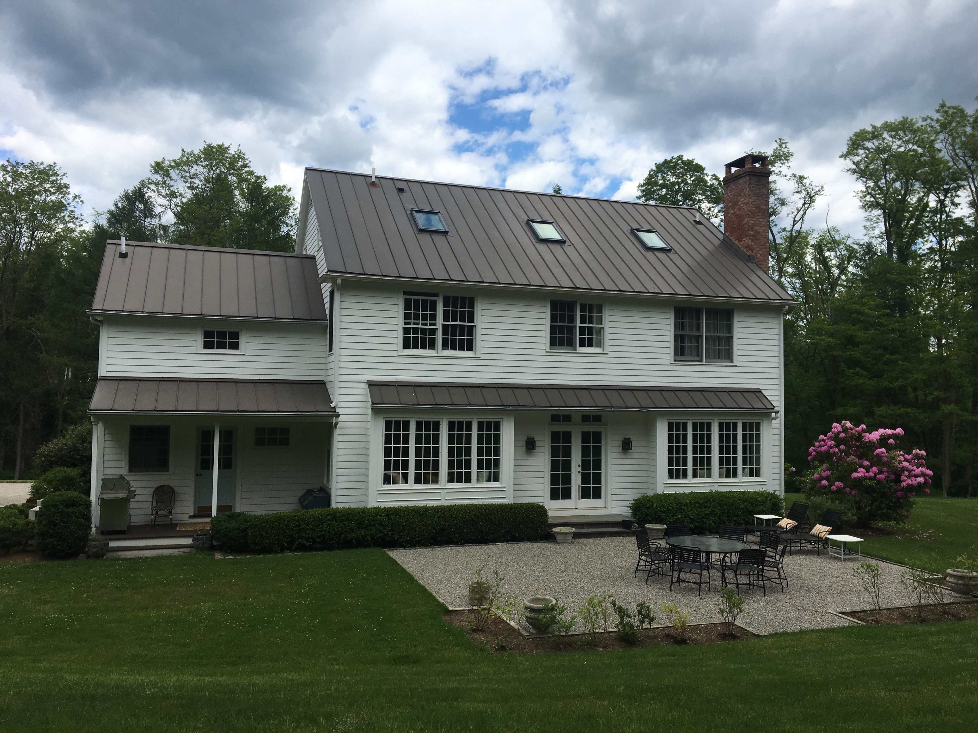 A white two-story house with a metal roof is surrounded by a lawn and garden, featuring a patio set and a flowering bush in the foreground.