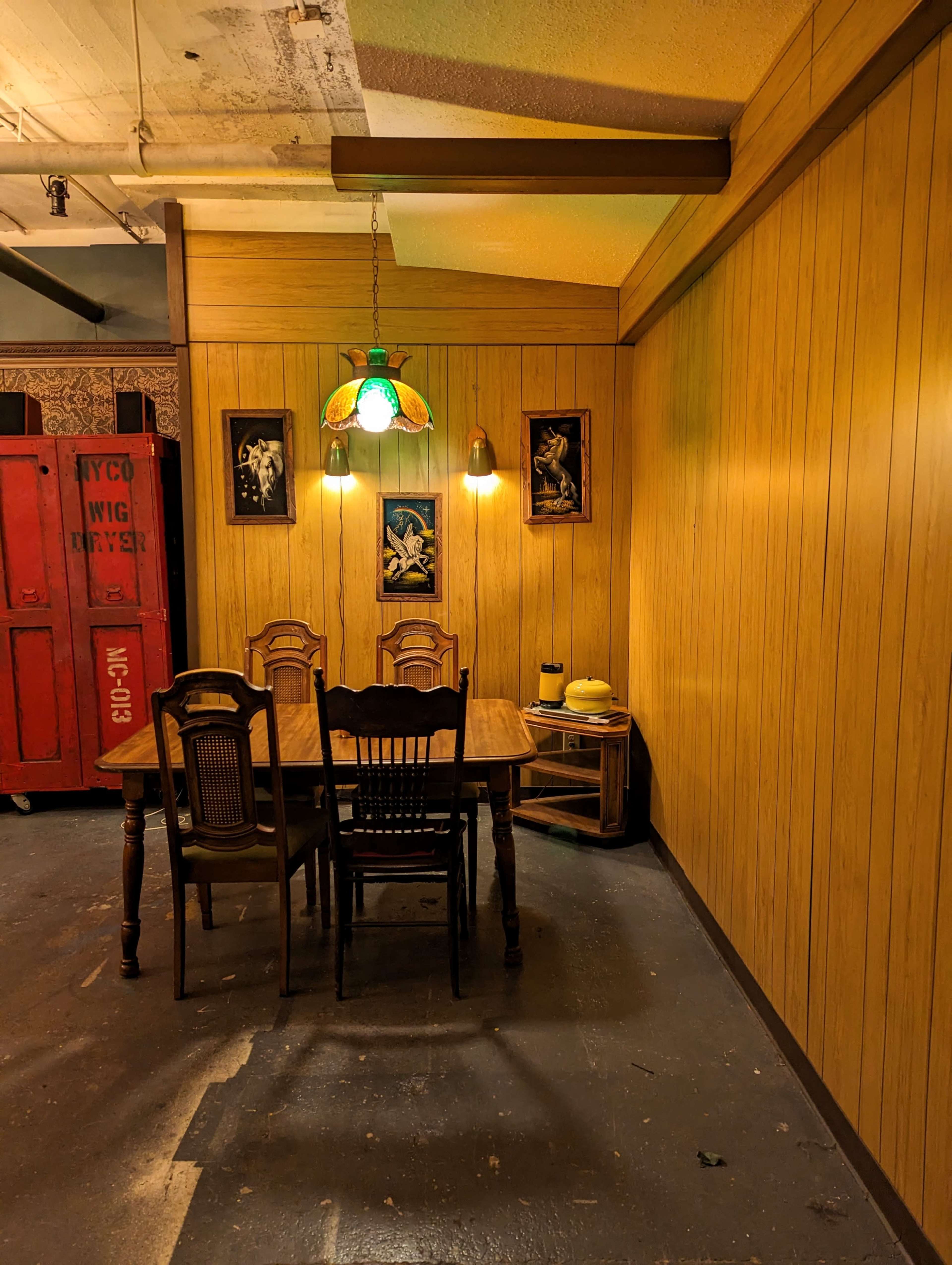 A wooden dining table with four chairs sits beneath a hanging lamp in a room featuring wooden walls and framed artworks.