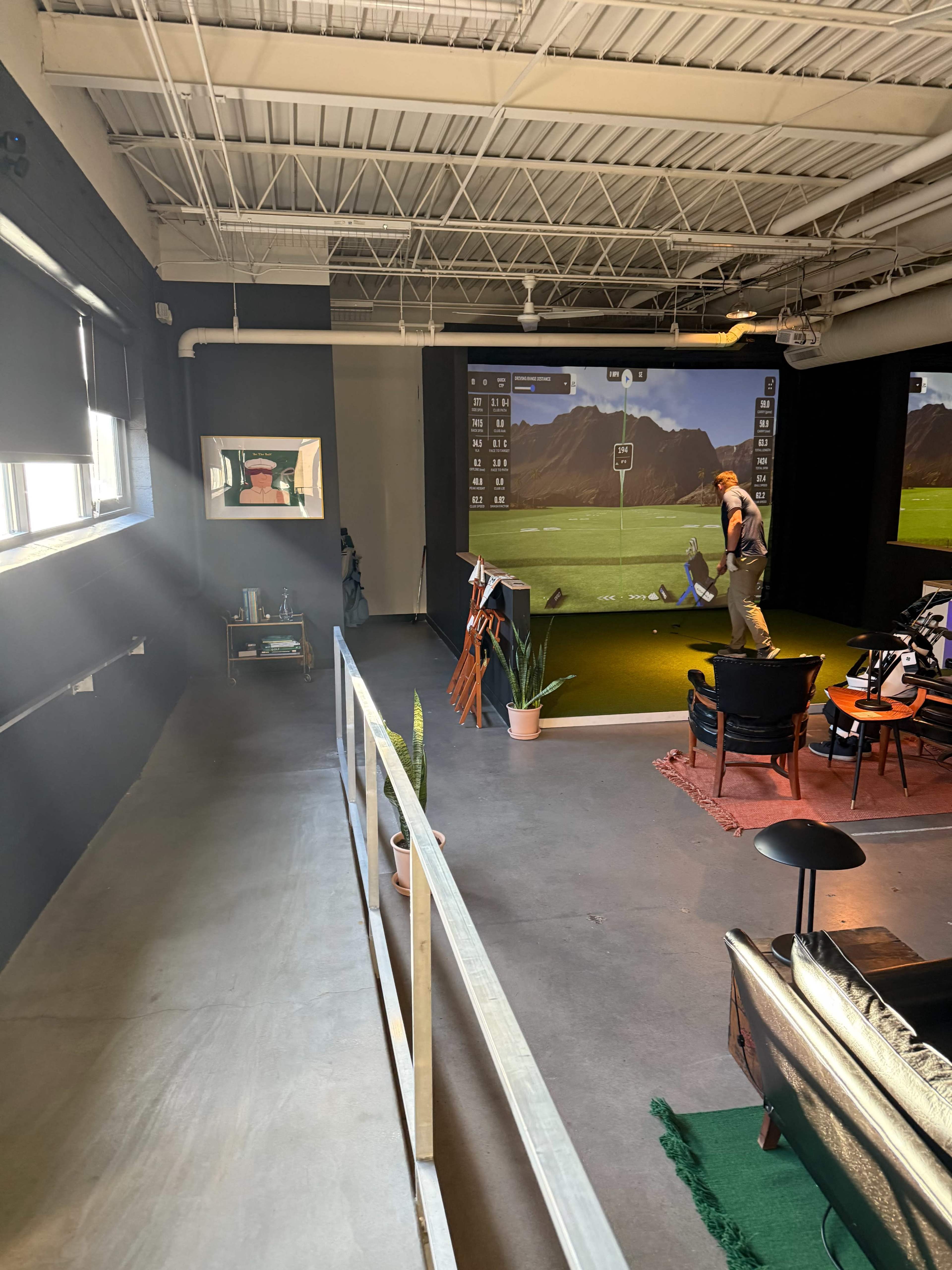 The image shows an indoor space designed for golf practice, featuring a putting green area with a virtual simulator, a person practicing golf, and various seating arrangements nearby.