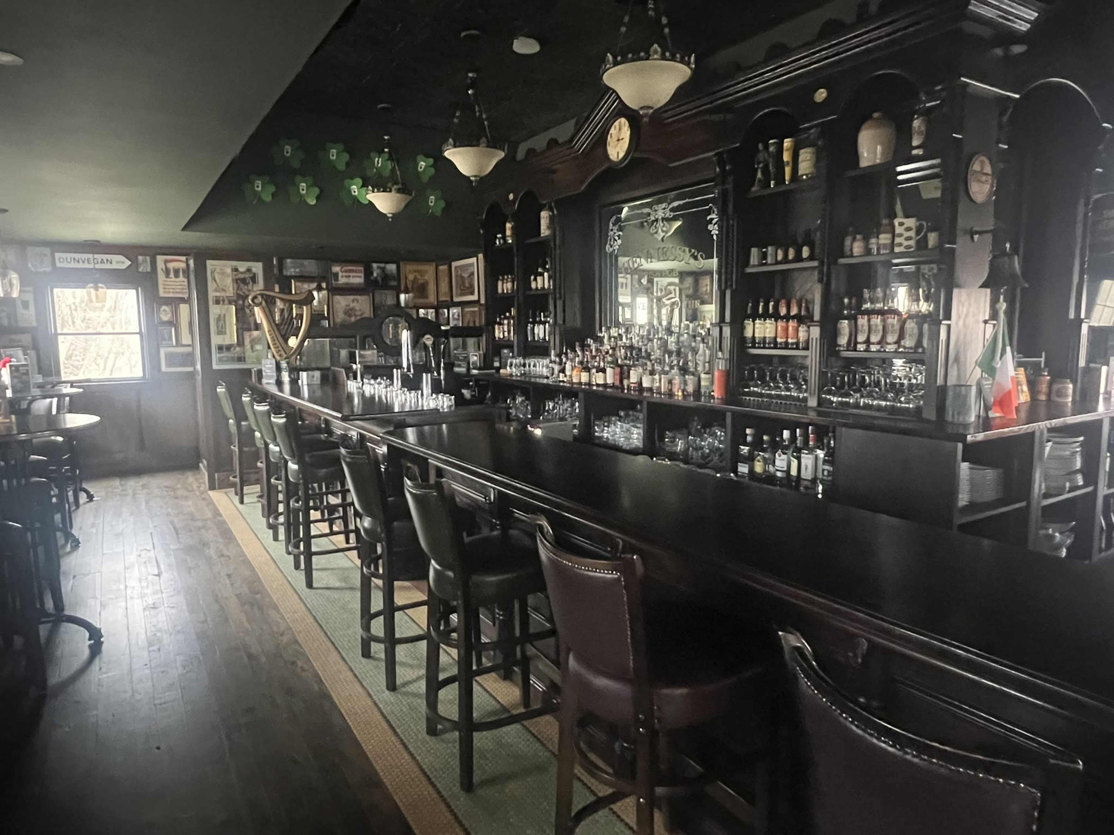 The image shows a polished wooden bar with high-backed chairs and shelves displaying various bottles of liquor in a dimly lit pub setting.