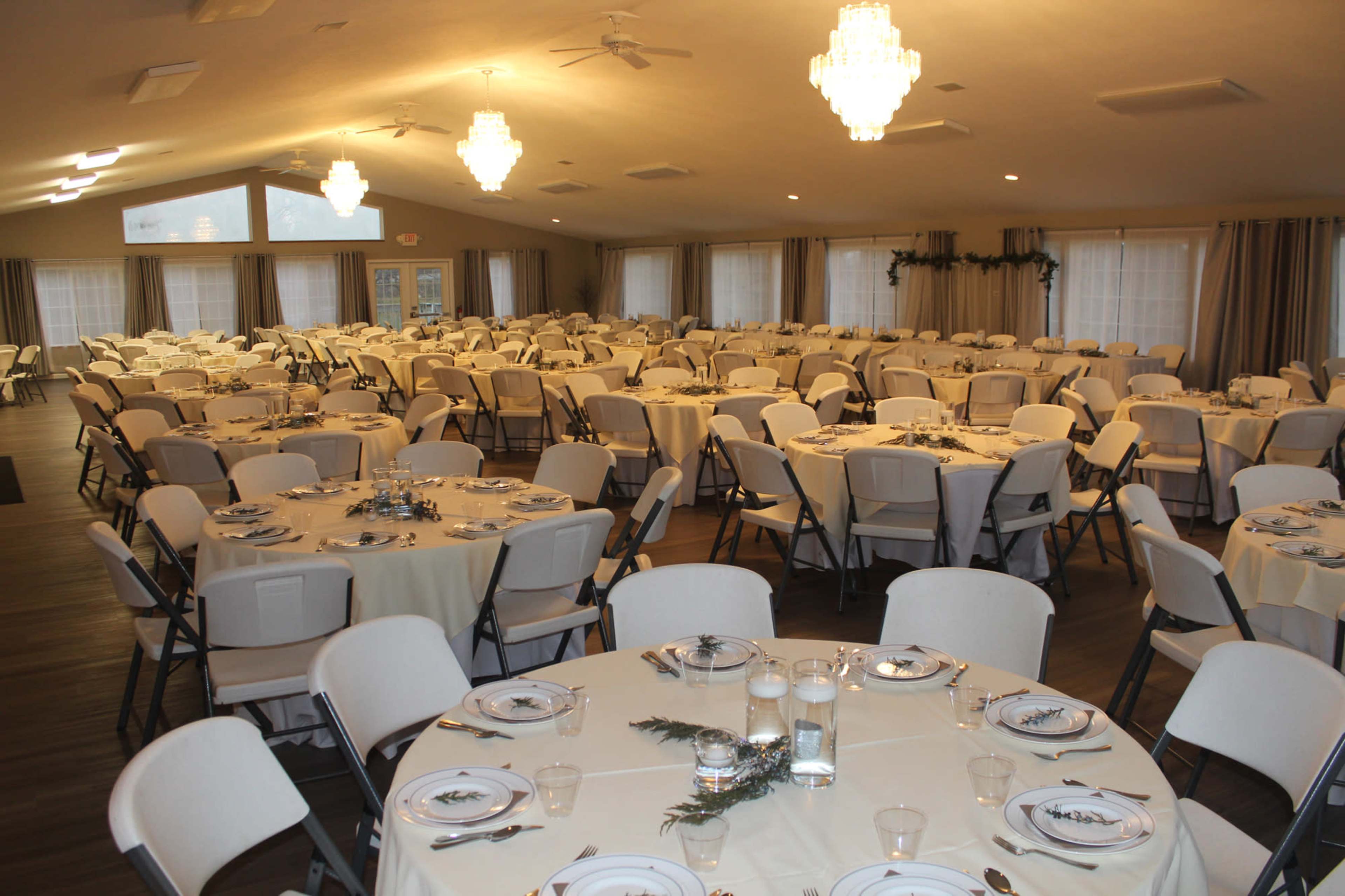 A large banquet hall set up with numerous round tables, each covered with white tablecloths and arranged with plates and glassware.