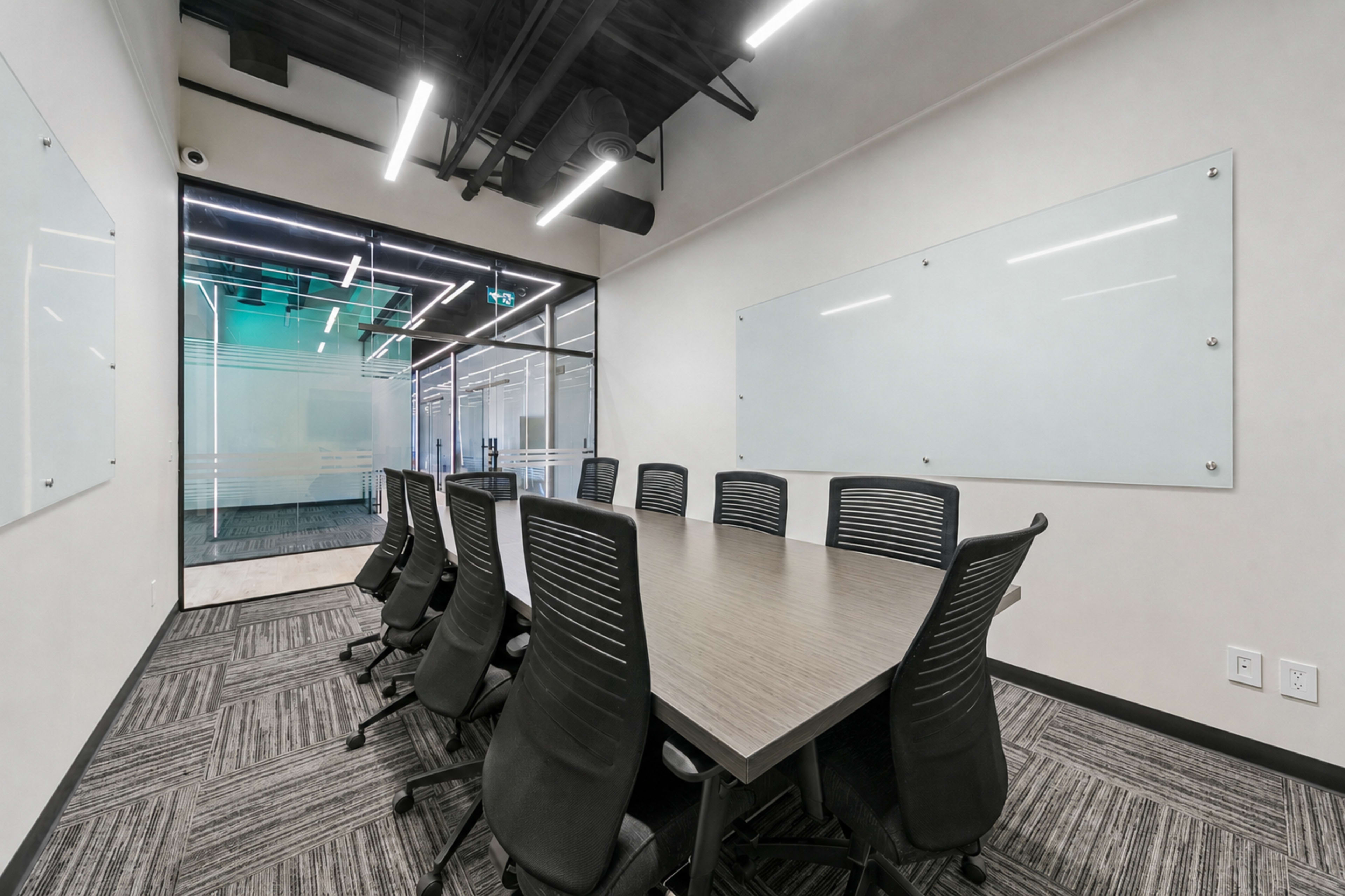 A modern conference room features a large rectangular table surrounded by black ergonomic chairs, with glass walls allowing visibility into an adjoining space.