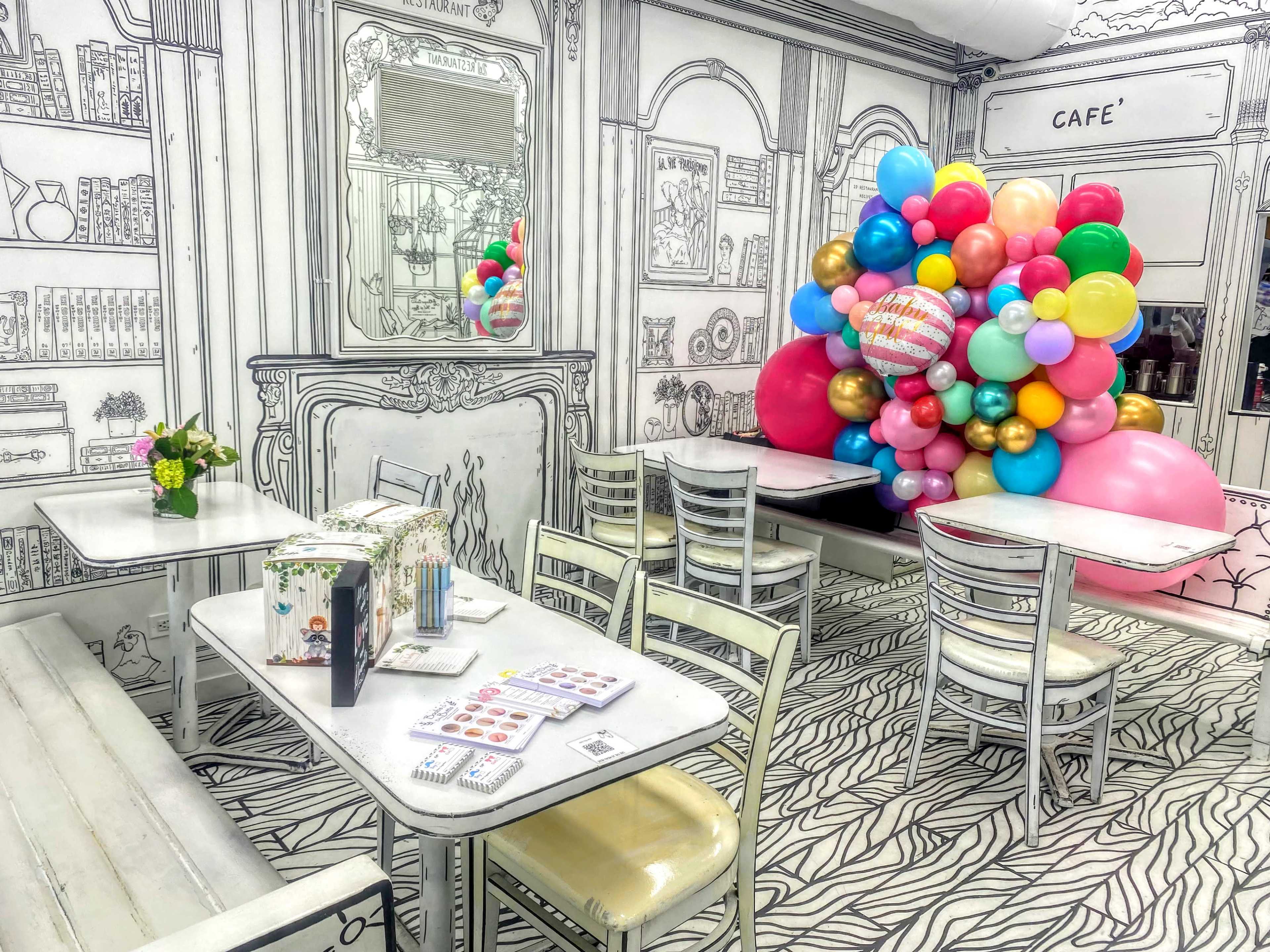 A whimsical café interior featuring outlined furniture and decorations, with colorful balloons arranged in a corner.