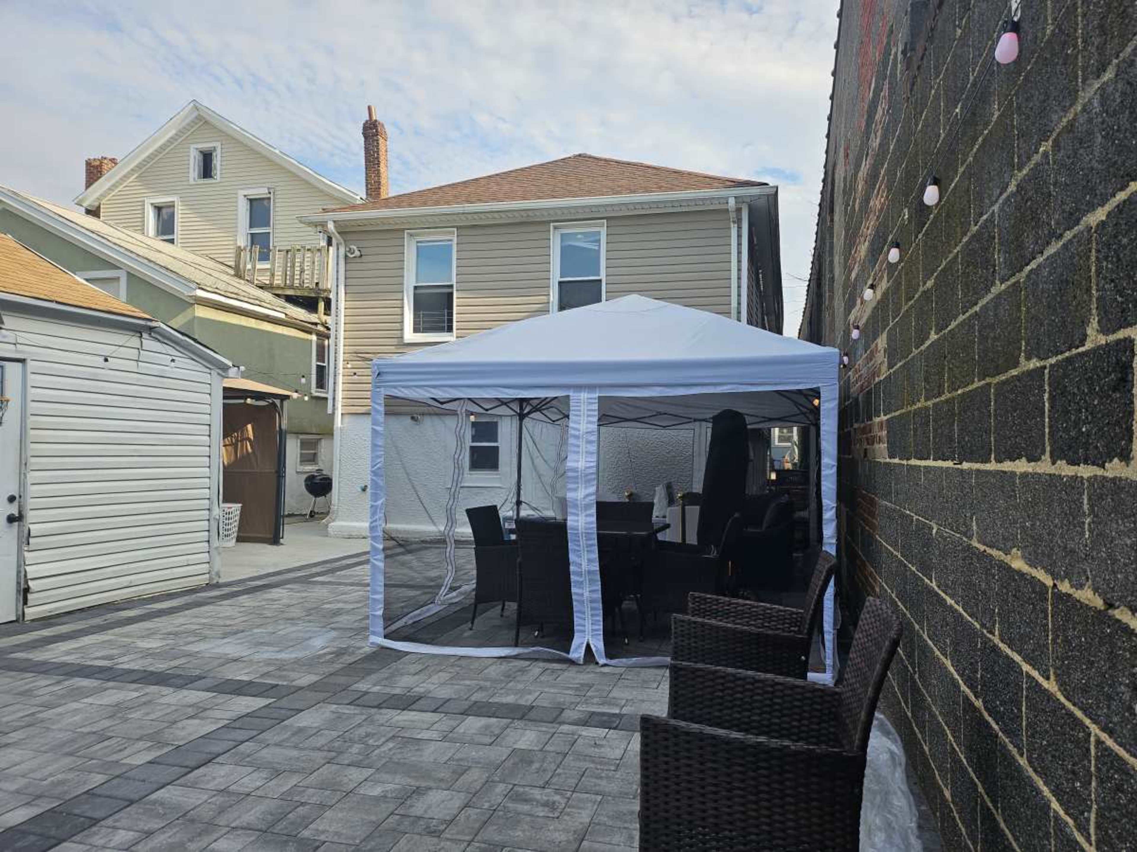 A white gazebo is set up on a paved patio next to a residential building, with outdoor furniture inside and various houses visible in the background.