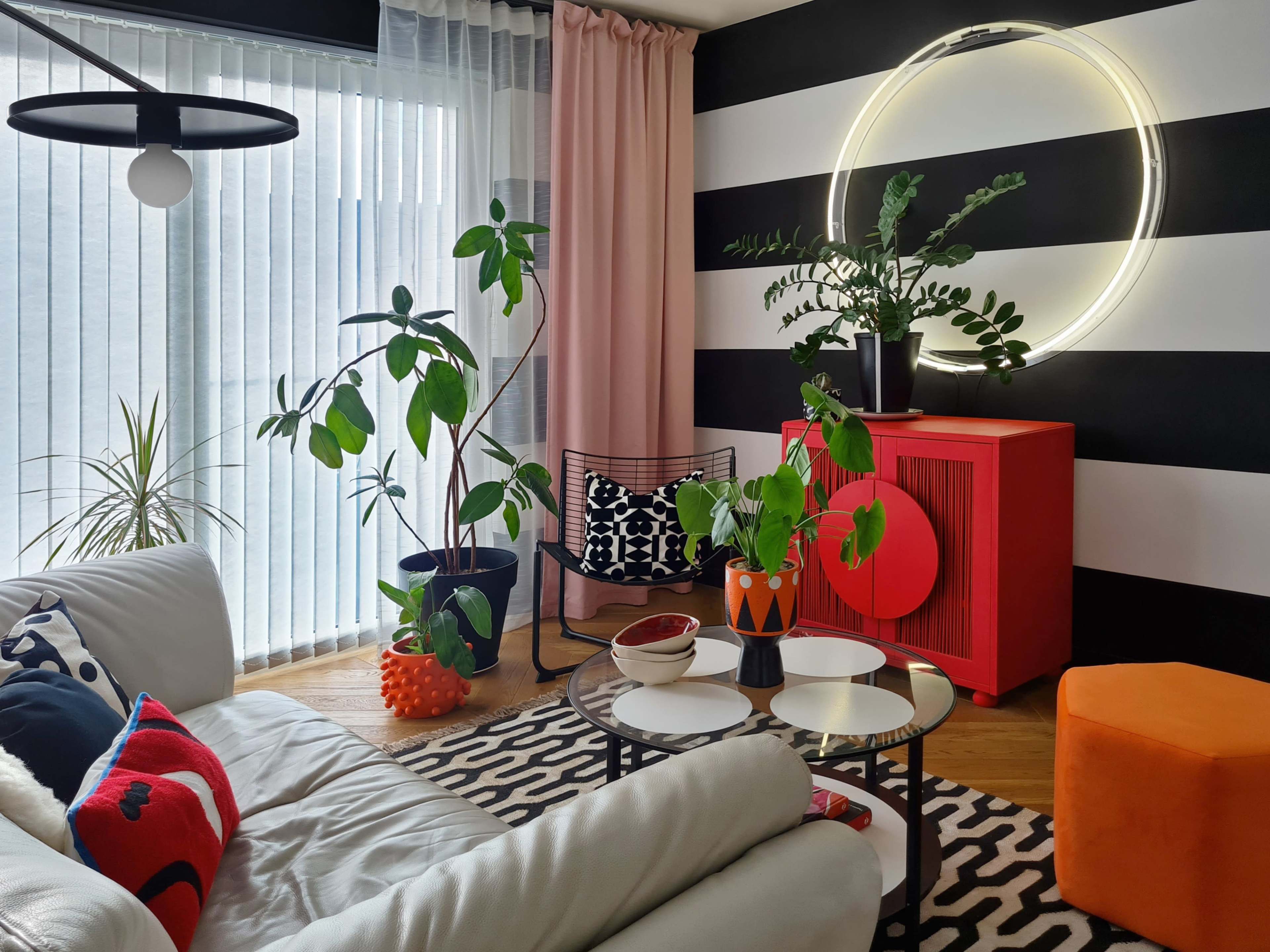 The living room features a white leather sofa, a round glass coffee table, and various indoor plants, set against a backdrop of black and white striped walls and a red cabinet.
