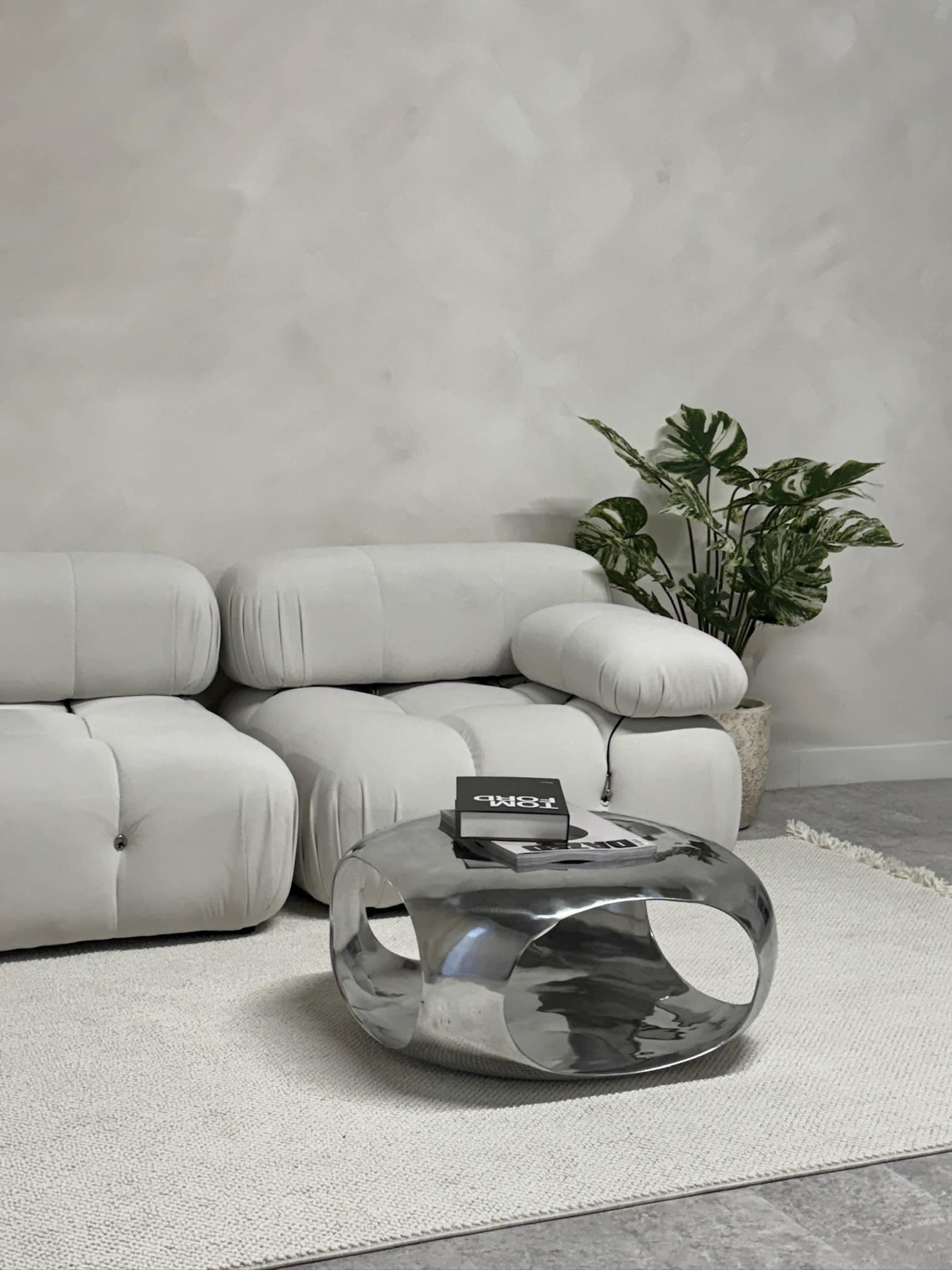 A light-colored, modern living room featuring a curved beige sofa next to a metallic coffee table and a potted plant against a textured wall.