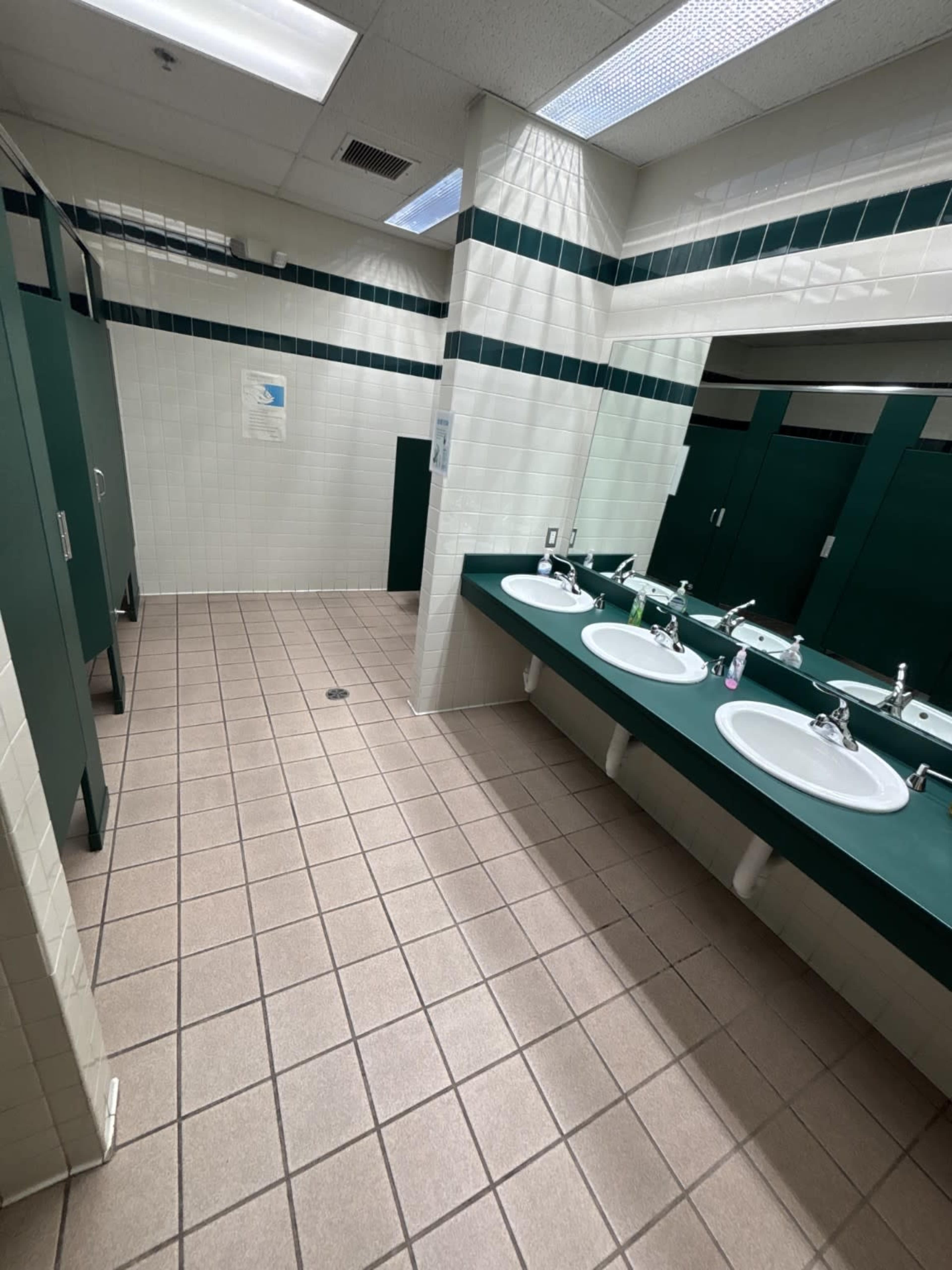 The image shows a public restroom with three sinks and separate stalls, featuring tiled floors and green accents.
