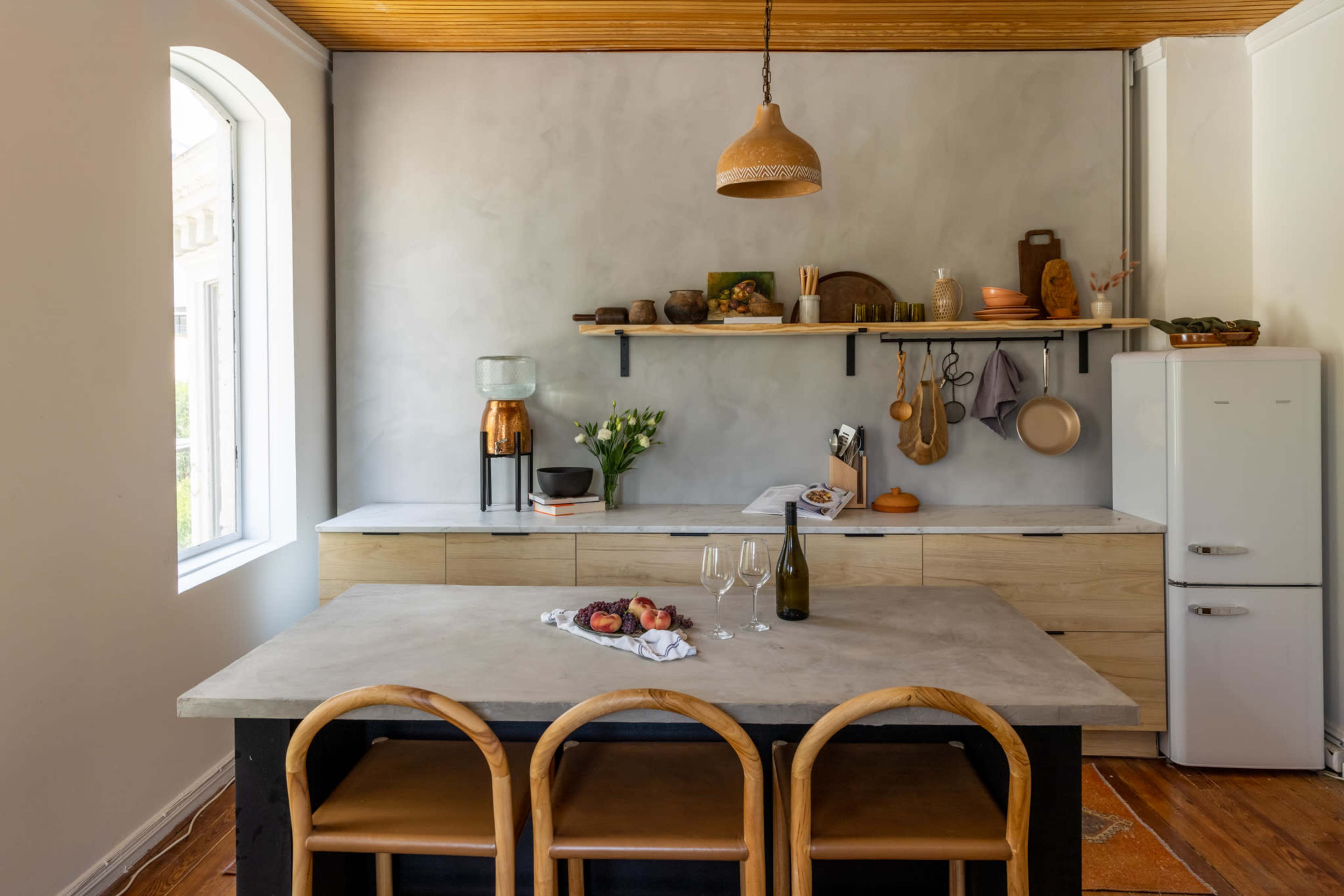 A modern kitchen features a concrete island with wooden bar stools, a vintage refrigerator, and a mix of cookware displayed on a wall shelf.
