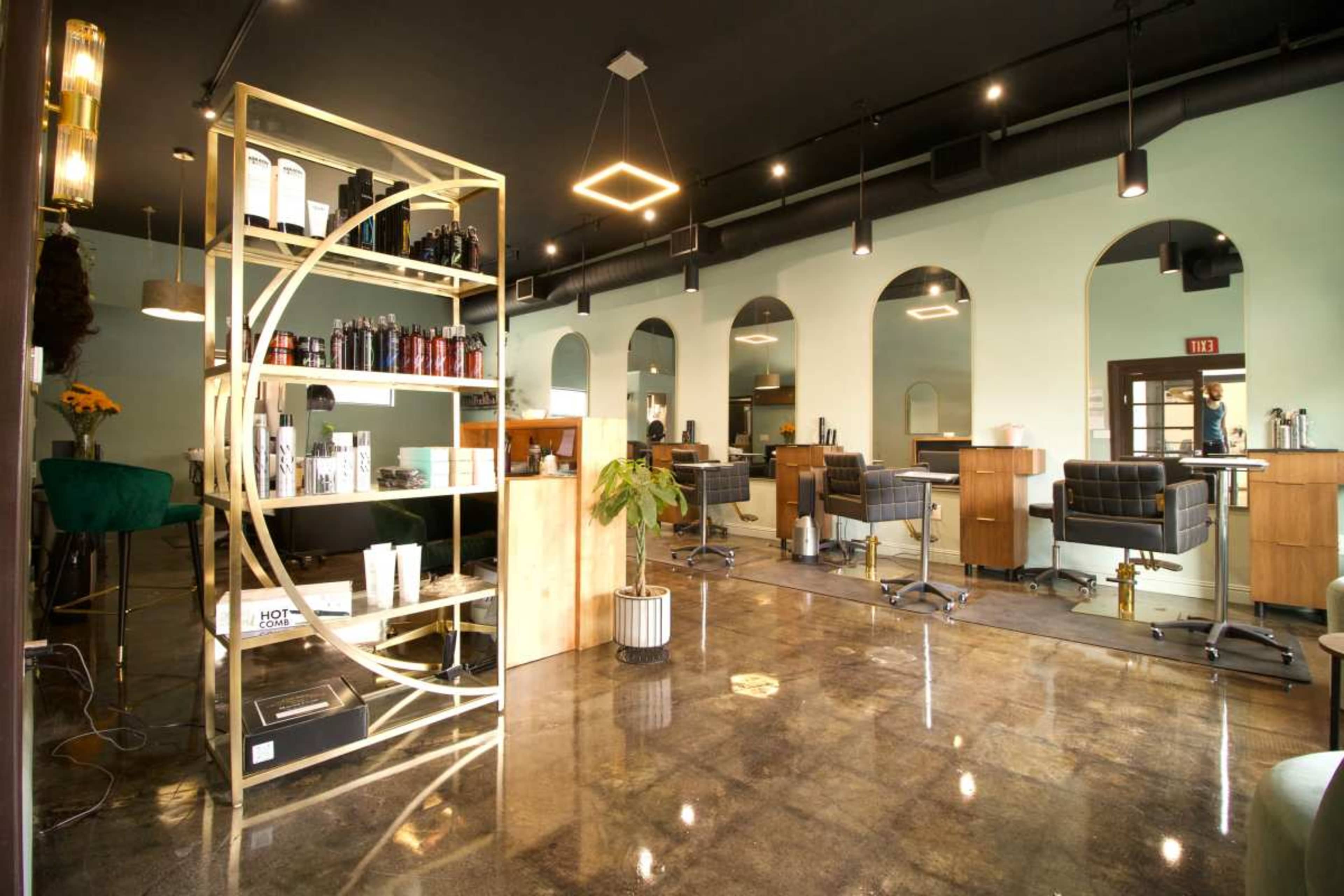 The image shows a modern hair salon space featuring sleek styling stations, decorative shelving, and a bright, airy atmosphere with green walls and polished flooring.