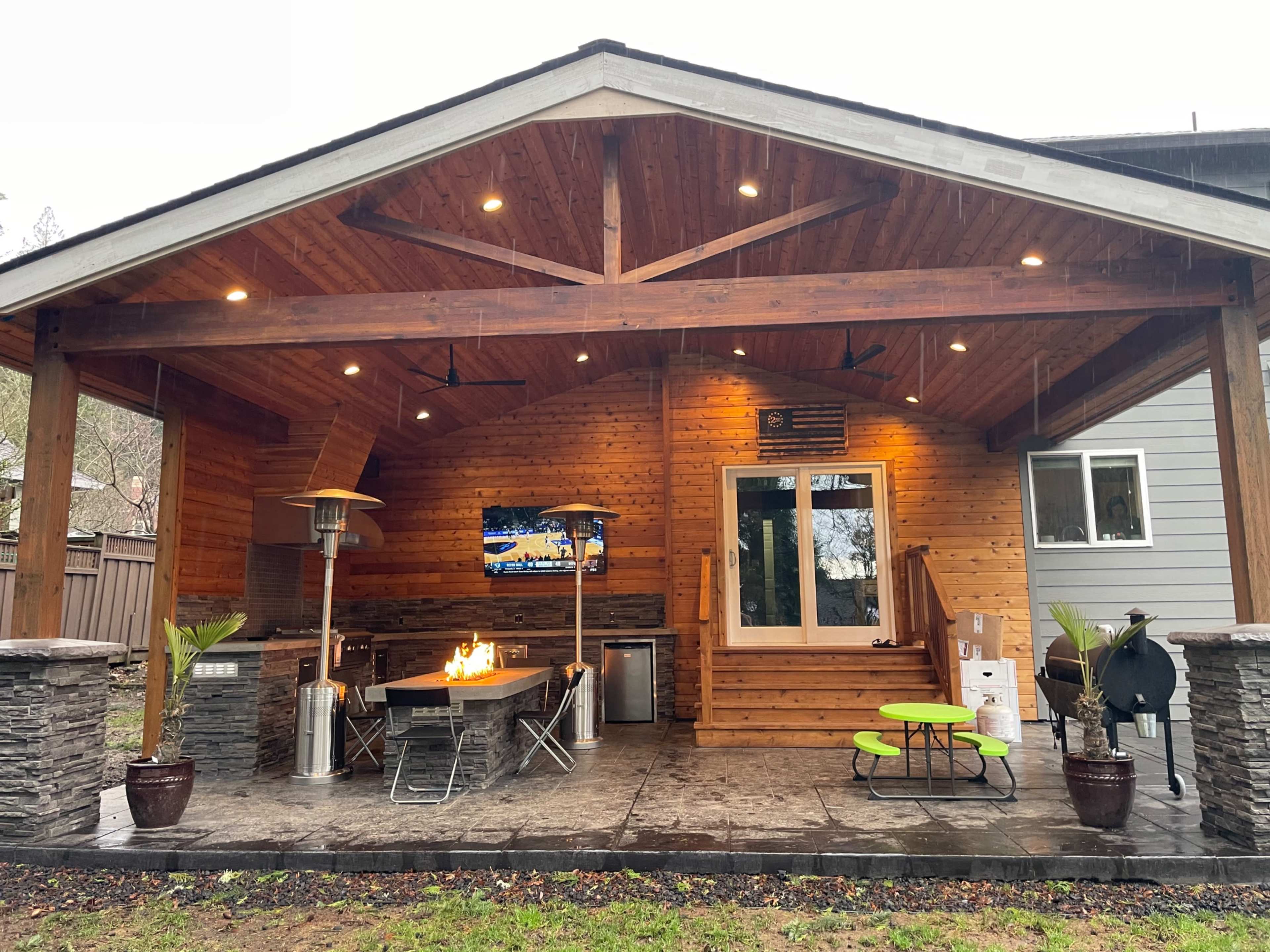 A covered outdoor patio featuring a grill, a dining table with chairs, a fire pit, and a television mounted on the wall, set against a wooden backdrop.