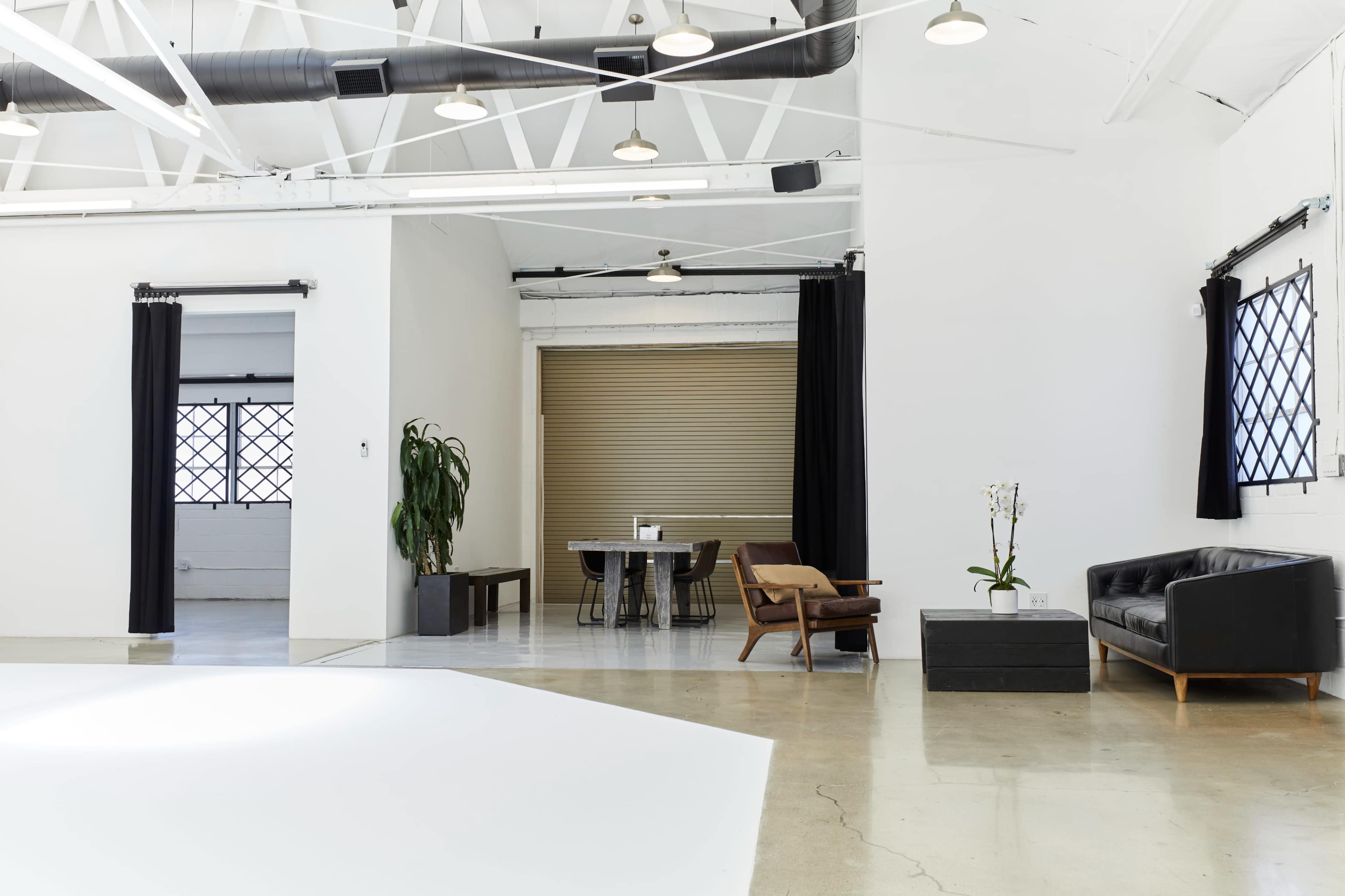 A spacious, modern interior features a black leather sofa, a wooden dining table, and large windows with dark curtains.