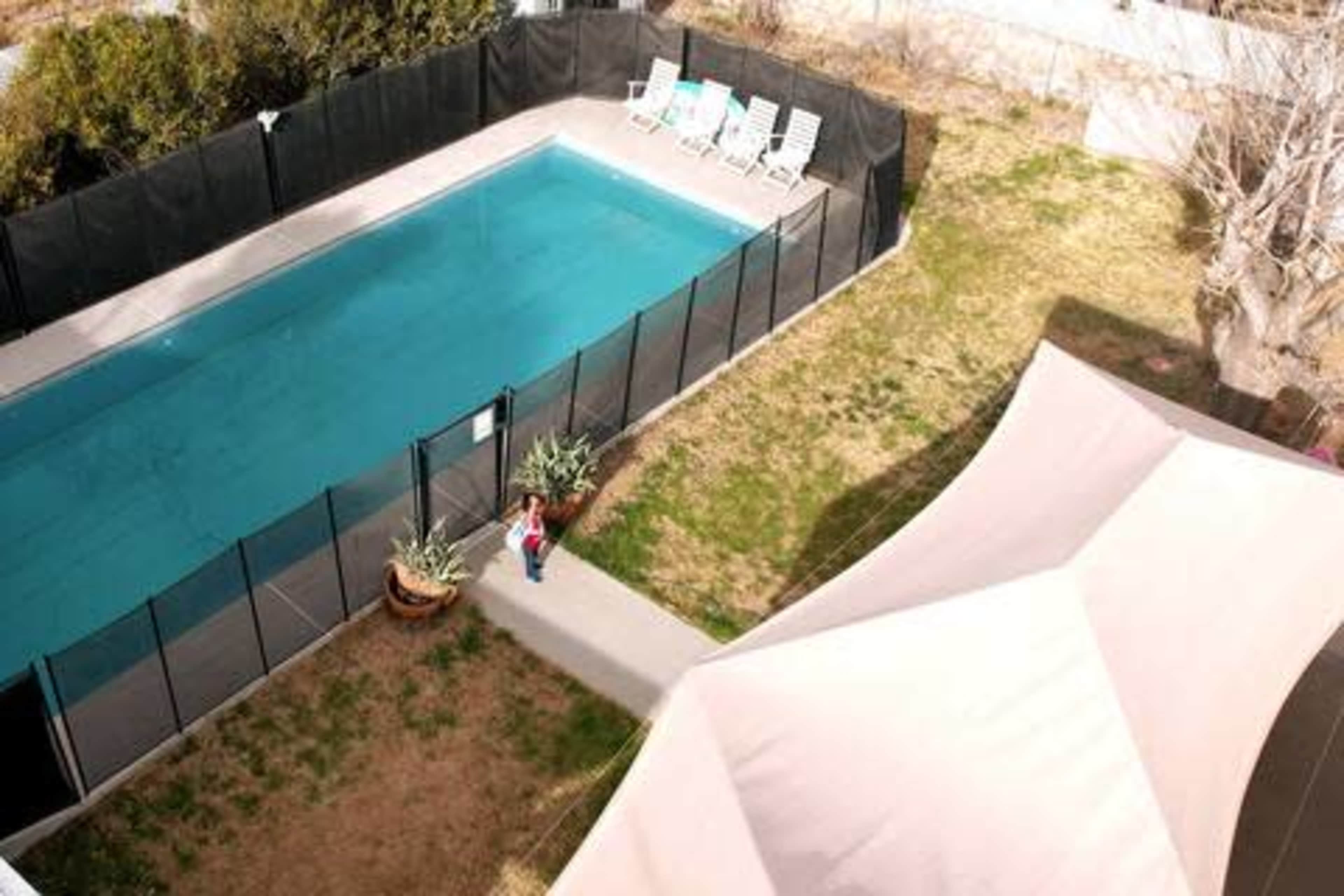 A rectangular swimming pool is enclosed by a black safety fence, with lounge chairs positioned nearby and a pathway leading to a person standing at the edge of the pool area.