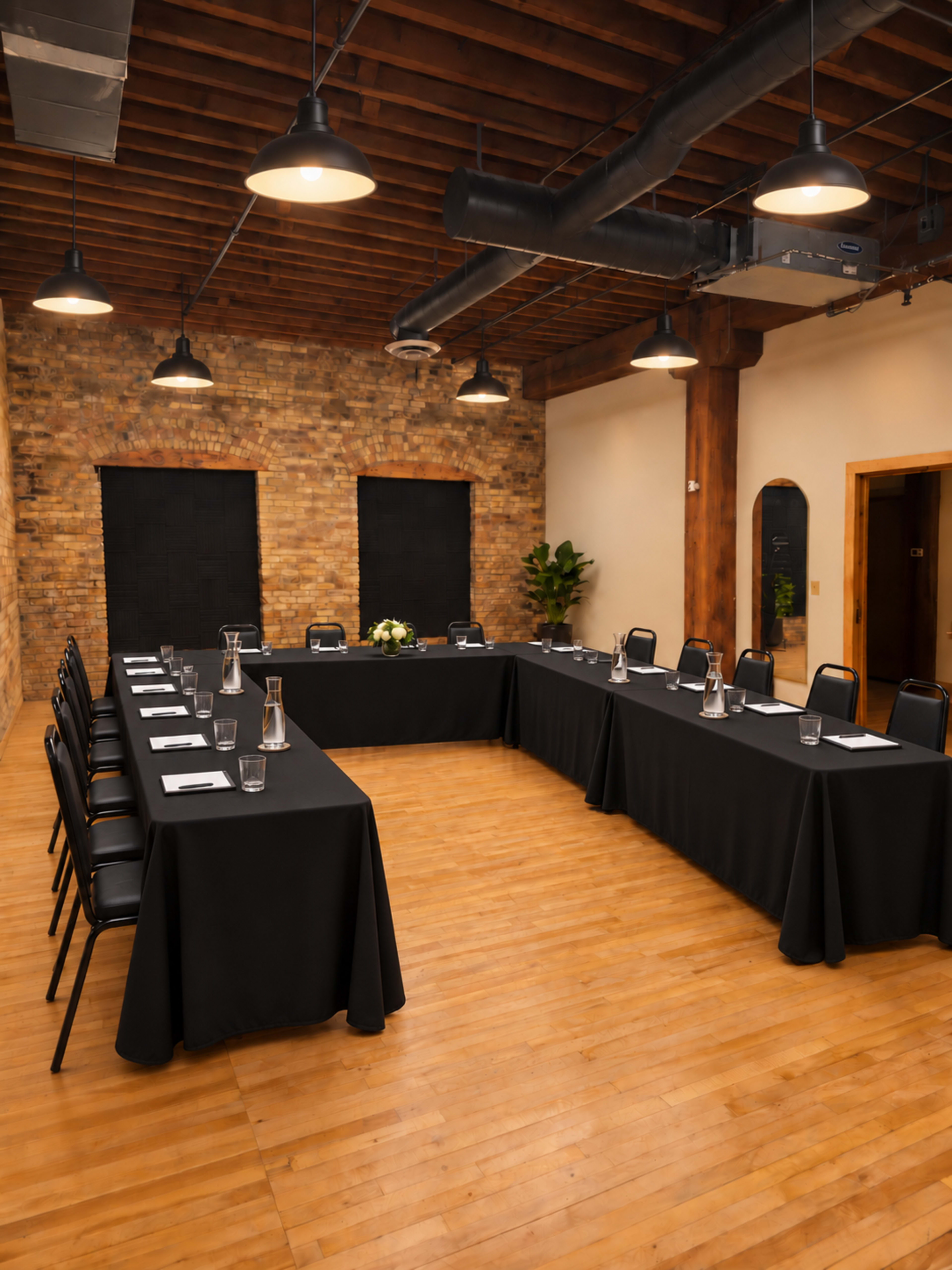 A spacious meeting room features two rectangular tables arranged in a U-shape, surrounded by black chairs, with exposed wooden beams and brick walls.