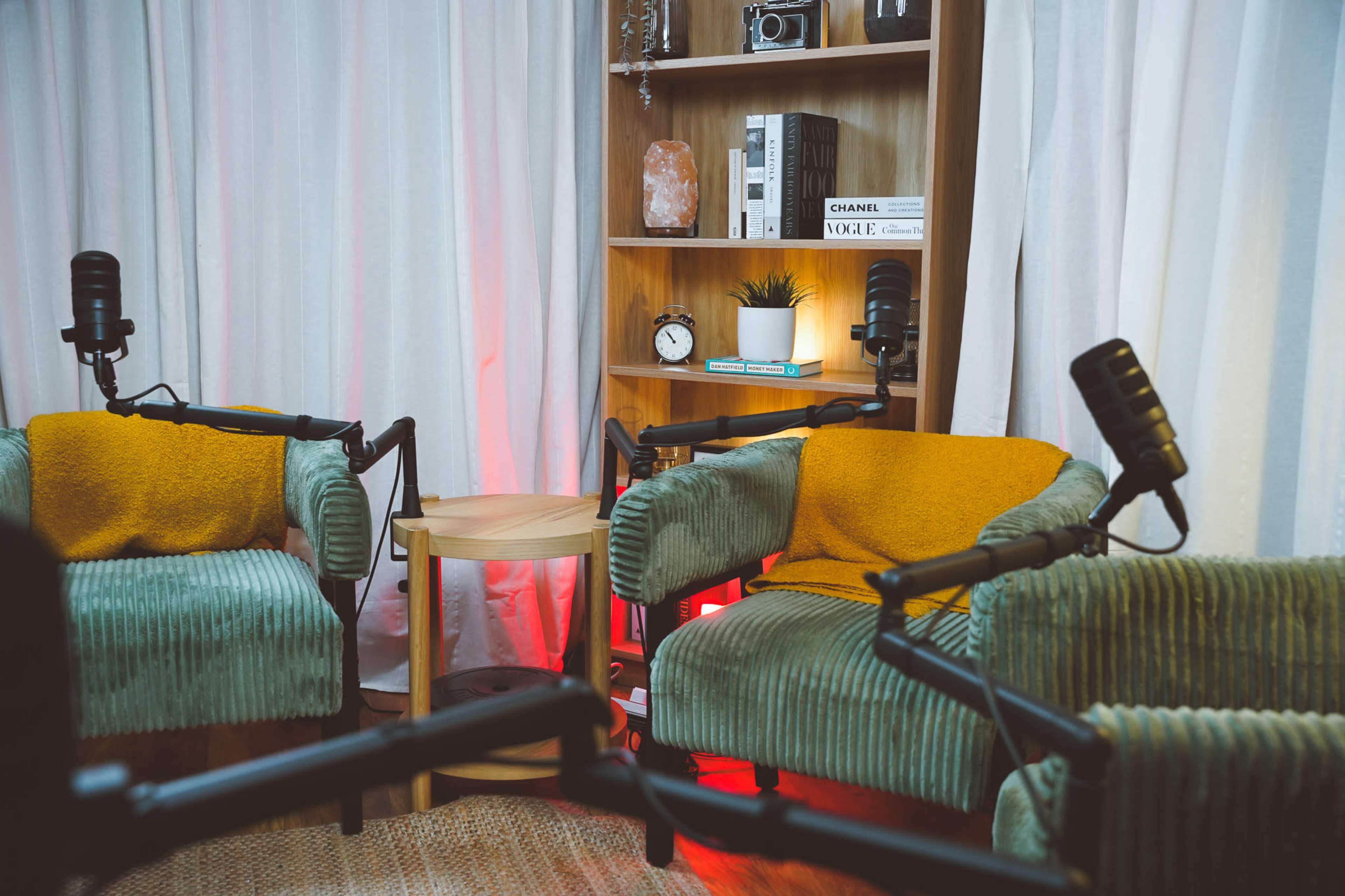 Two green armchairs with orange throws are positioned in a cozy setting, featuring a small side table, microphones, and a shelf with decor and books in the background.