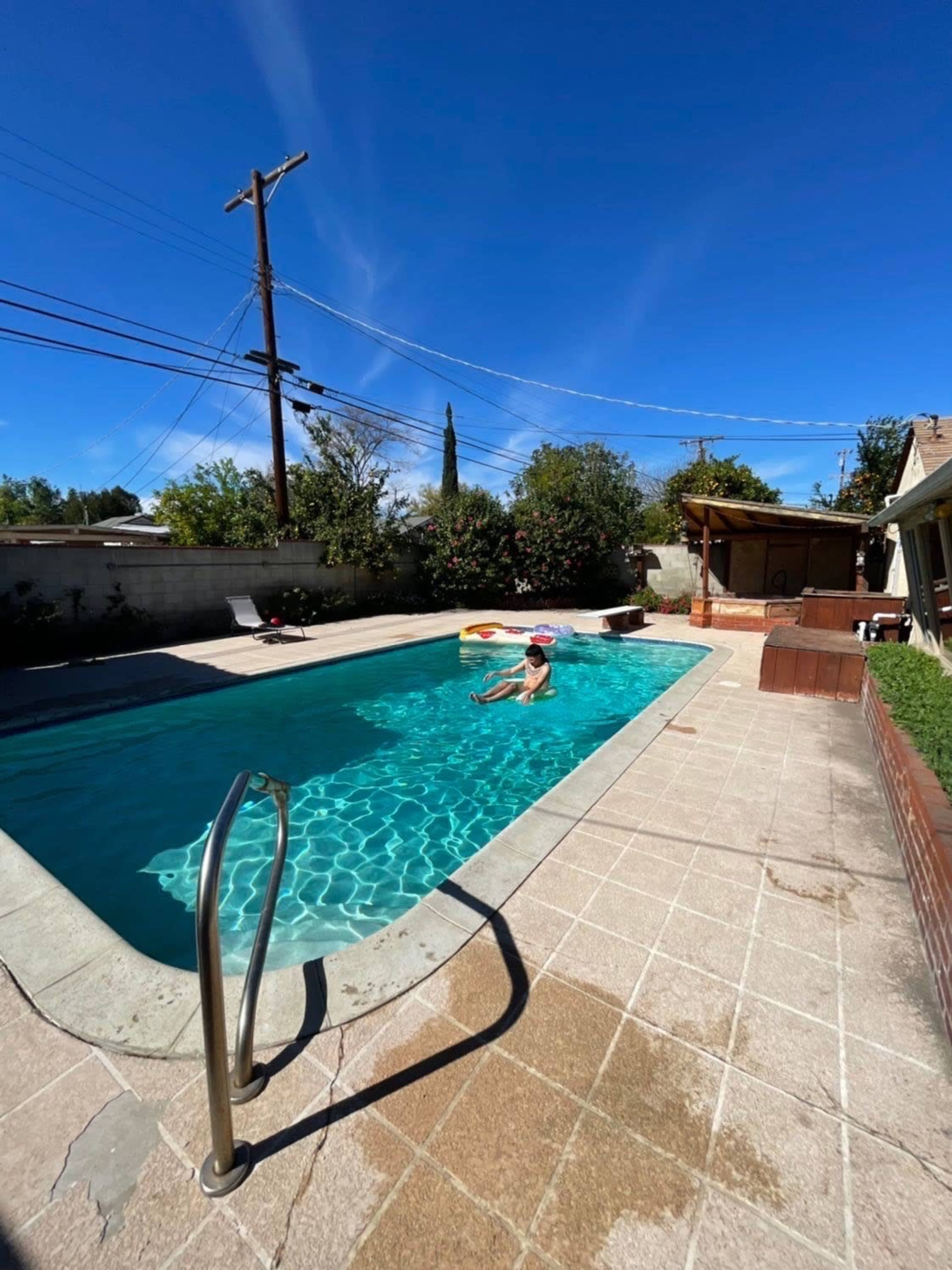 A person is swimming in a residential pool surrounded by trees and a wooden structure on the patio.