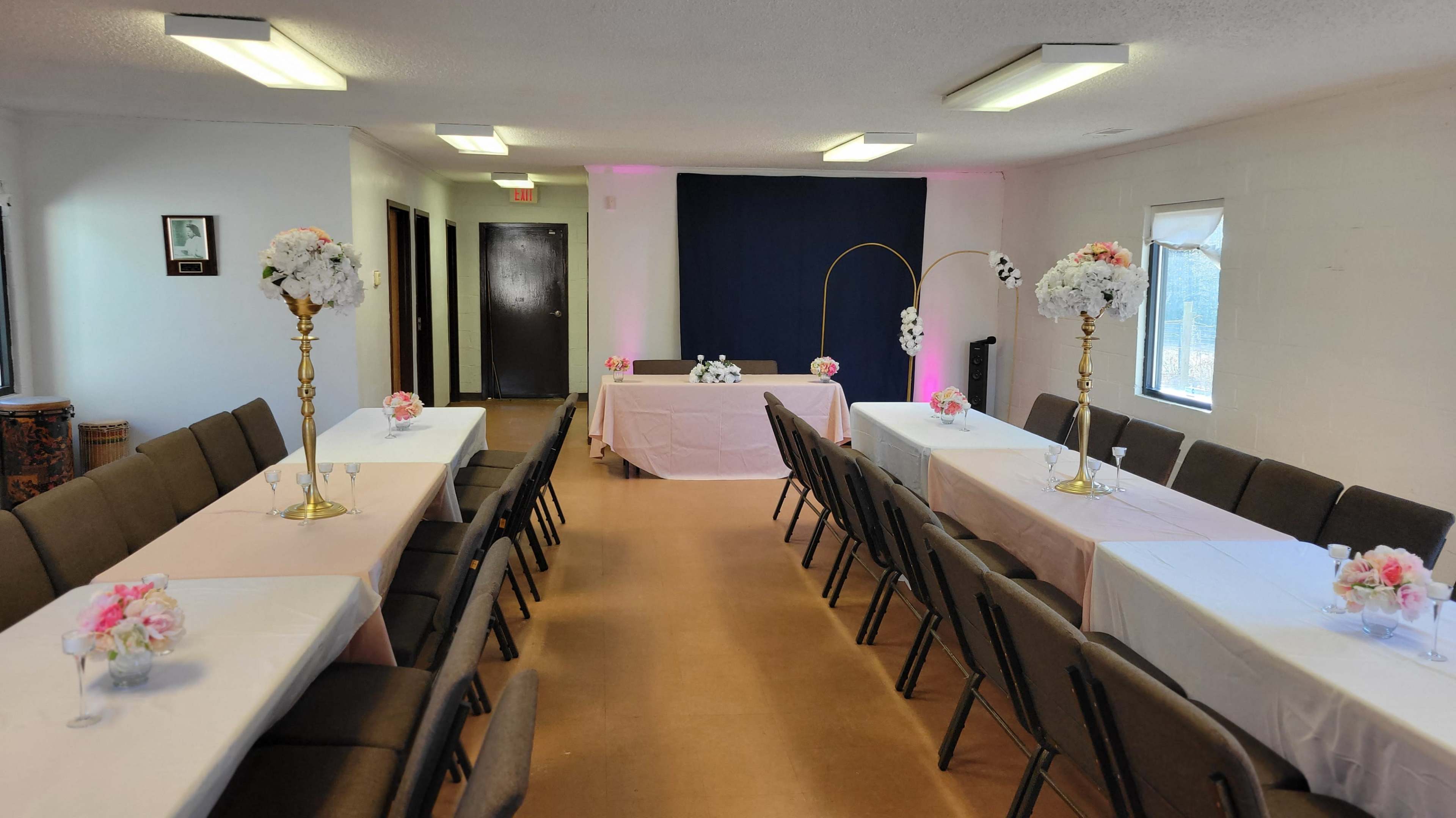 The image shows a decorated event space with two rows of tables set with floral arrangements, leading to a backdrop for a main display.