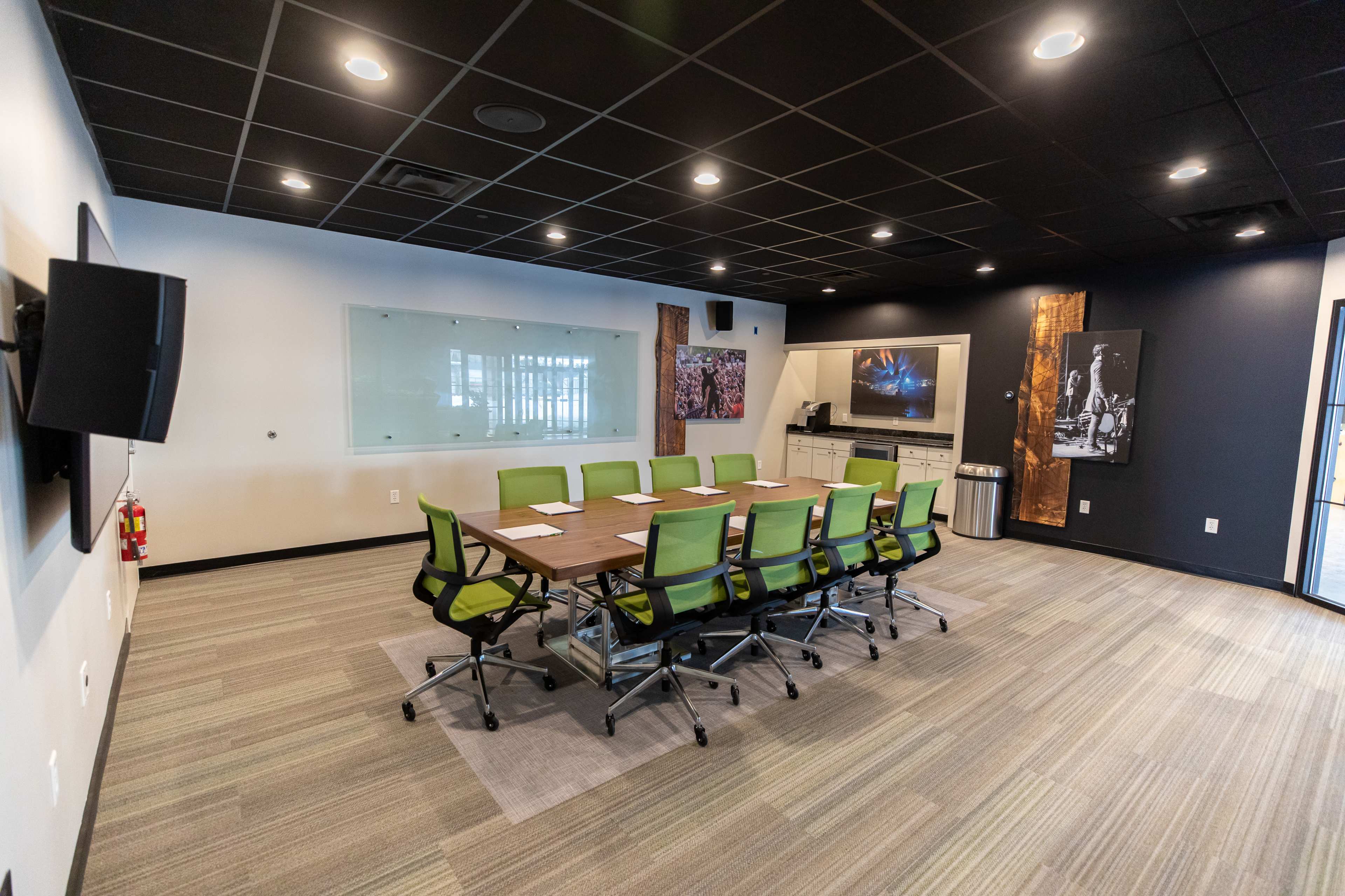 A modern conference room features a large rectangular table surrounded by green swivel chairs, with a glass wall and wall-mounted screens displaying artwork.