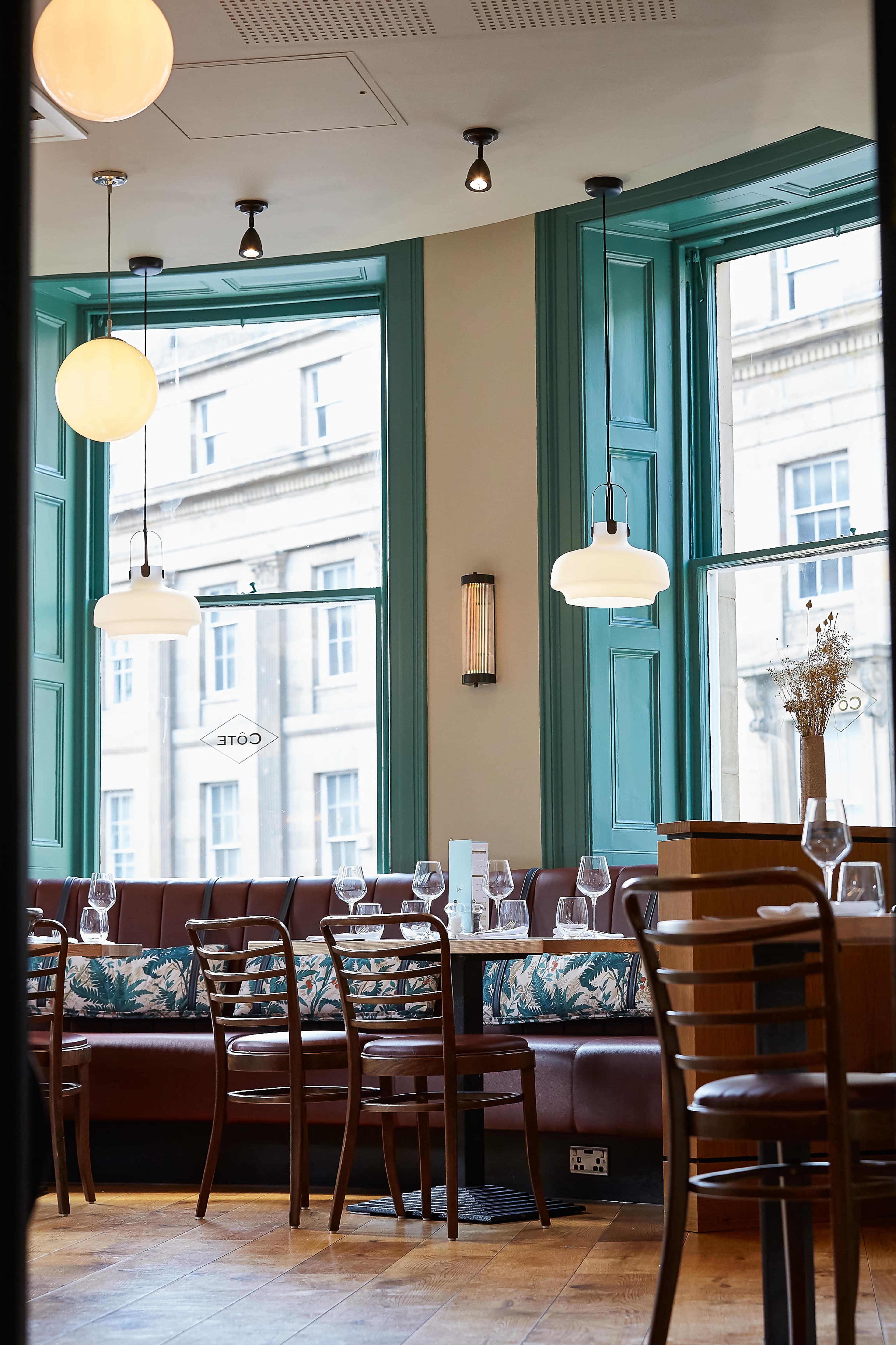 The image shows a restaurant interior featuring green windows, wooden chairs, and pendant lighting above a semi-circular booth with floral-patterned cushions.
