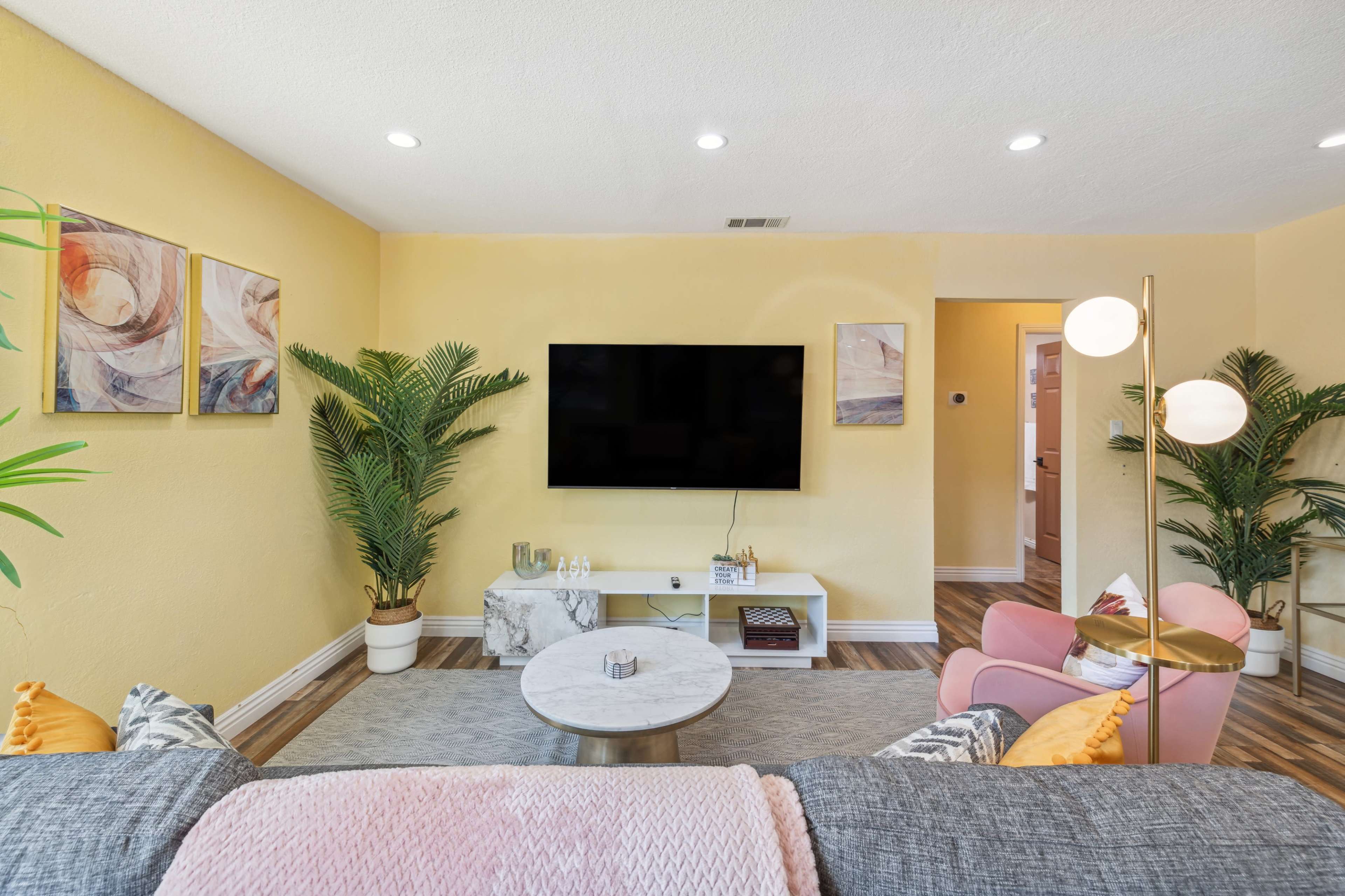 A bright living room features a large TV mounted on a yellow wall, a round coffee table, pink chairs, and potted plants.