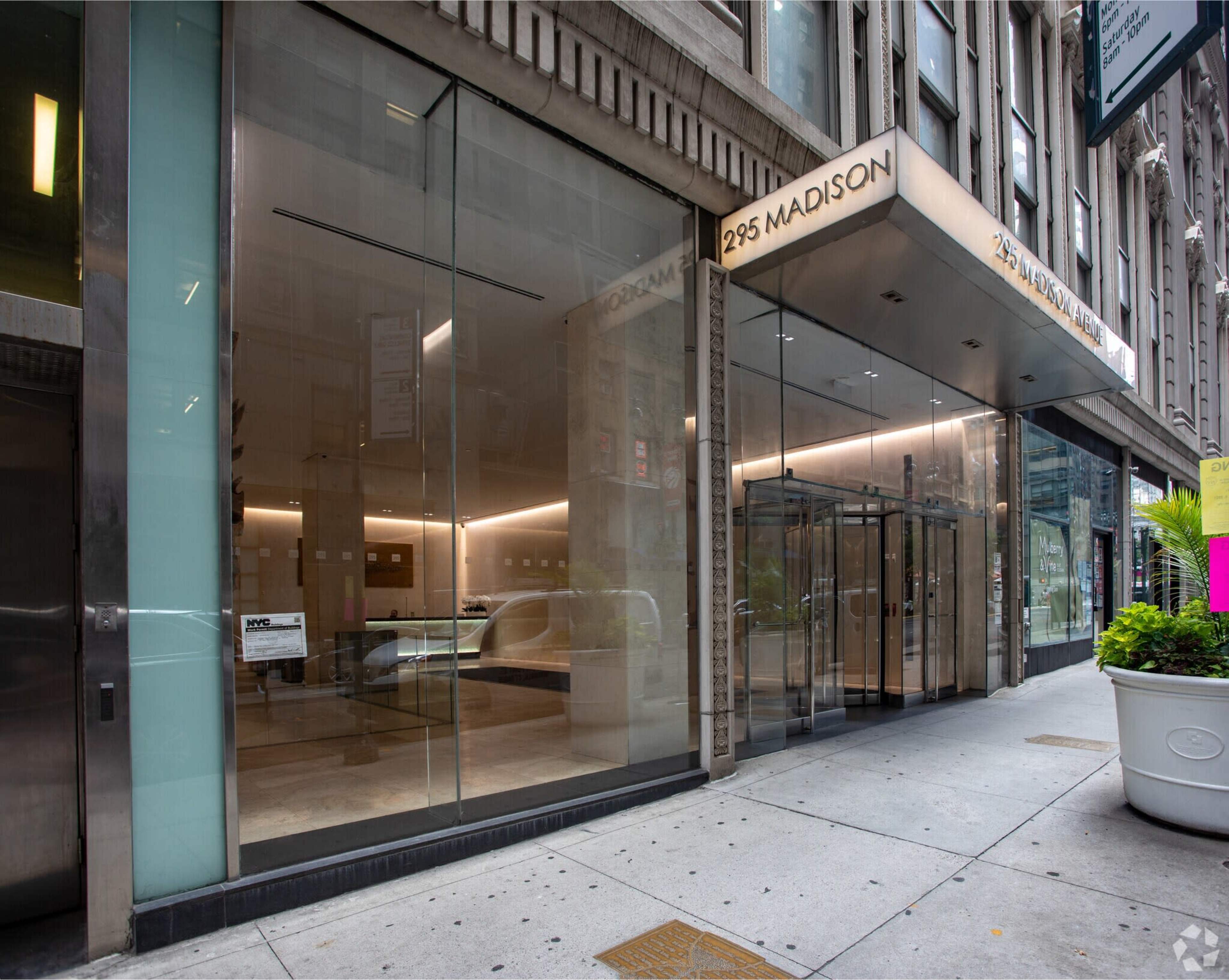 The image shows the modern glass entrance of a high-rise building located at 295 Madison Avenue, featuring clean lines and an open sidewalk.