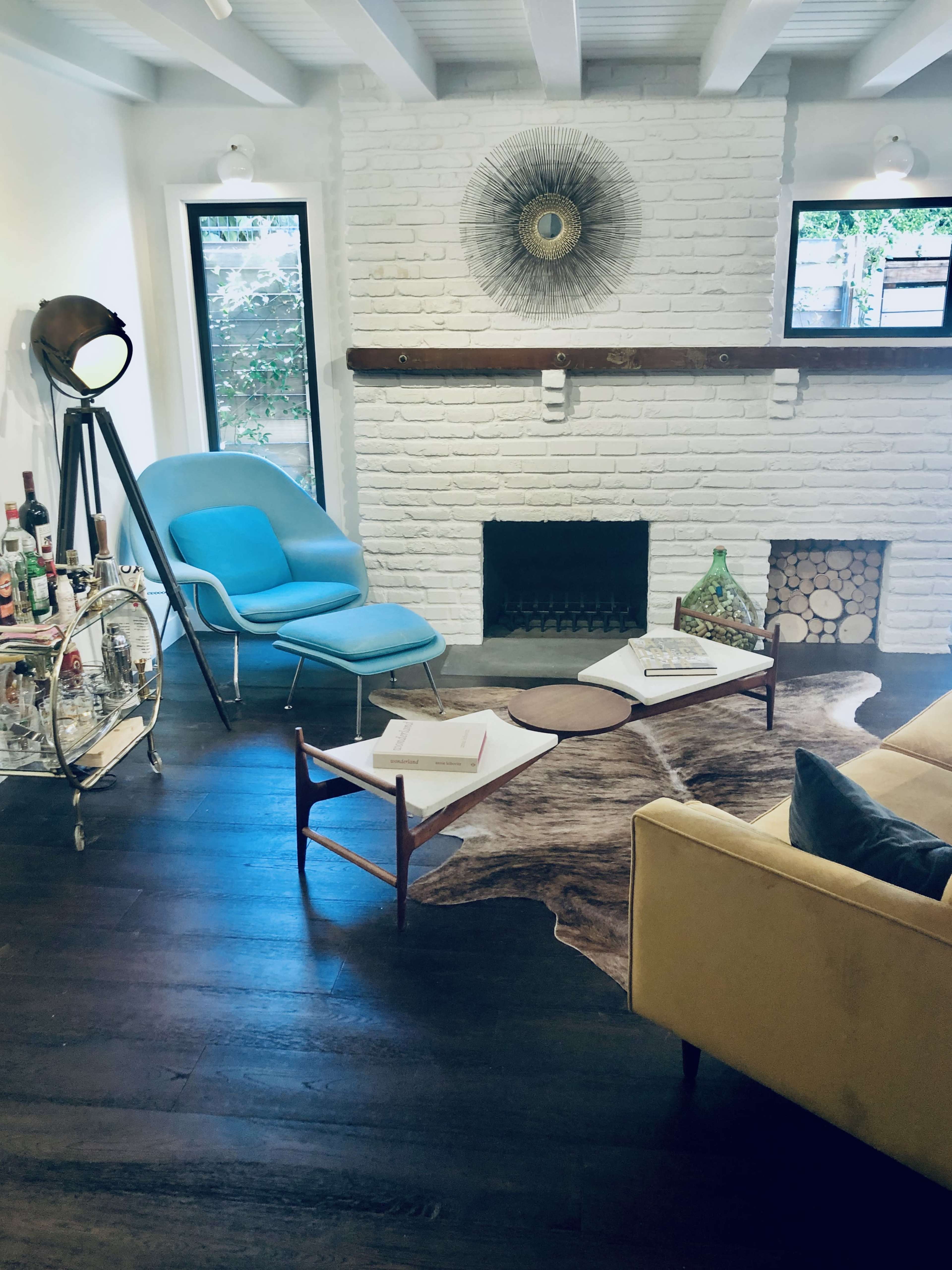 The image shows a stylish living room featuring a blue chair, a wooden coffee table, a vintage bar cart with bottles, and a fireplace, all set against white brick walls and dark wooden flooring.
