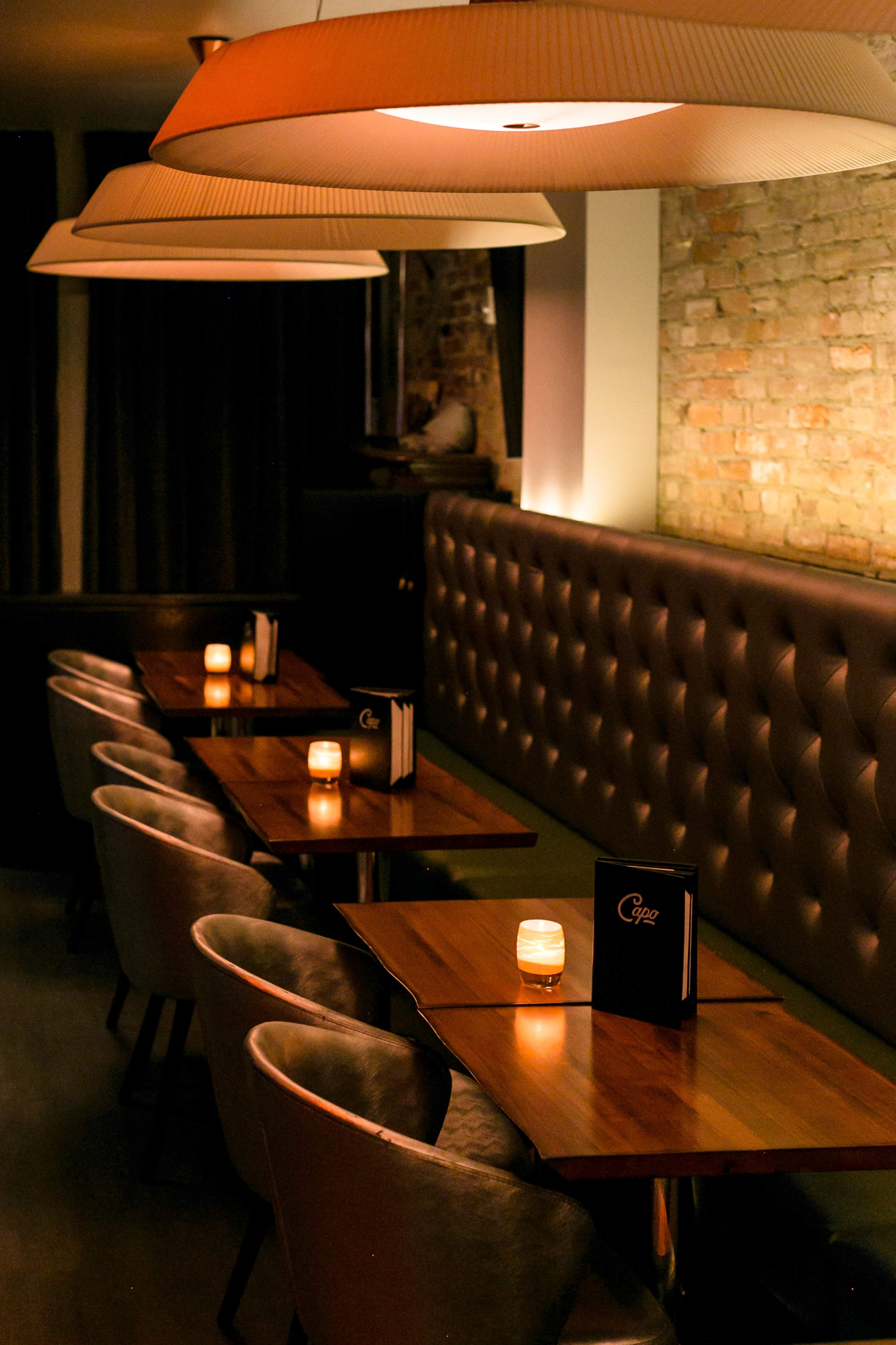 A dimly lit restaurant interior with wooden tables, plush chairs, and soft lighting from overhead lamps.
